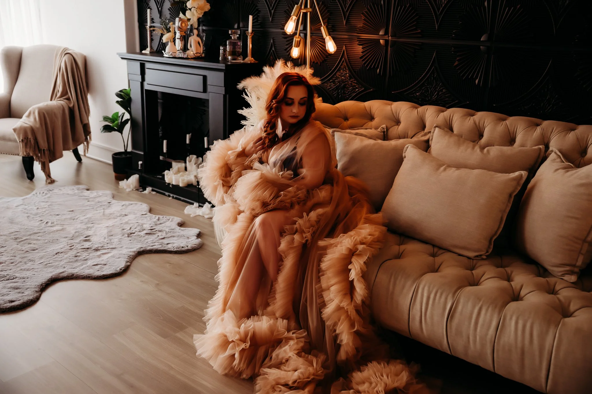 A woman in a flowing beige and ruffled dress sitting on a beige tufted sofa in a dimly lit, stylish living room with candles, candles, candles, and decorative items on a black fireplace mantel, a beige armchair, a white rug, and a black wall with geo