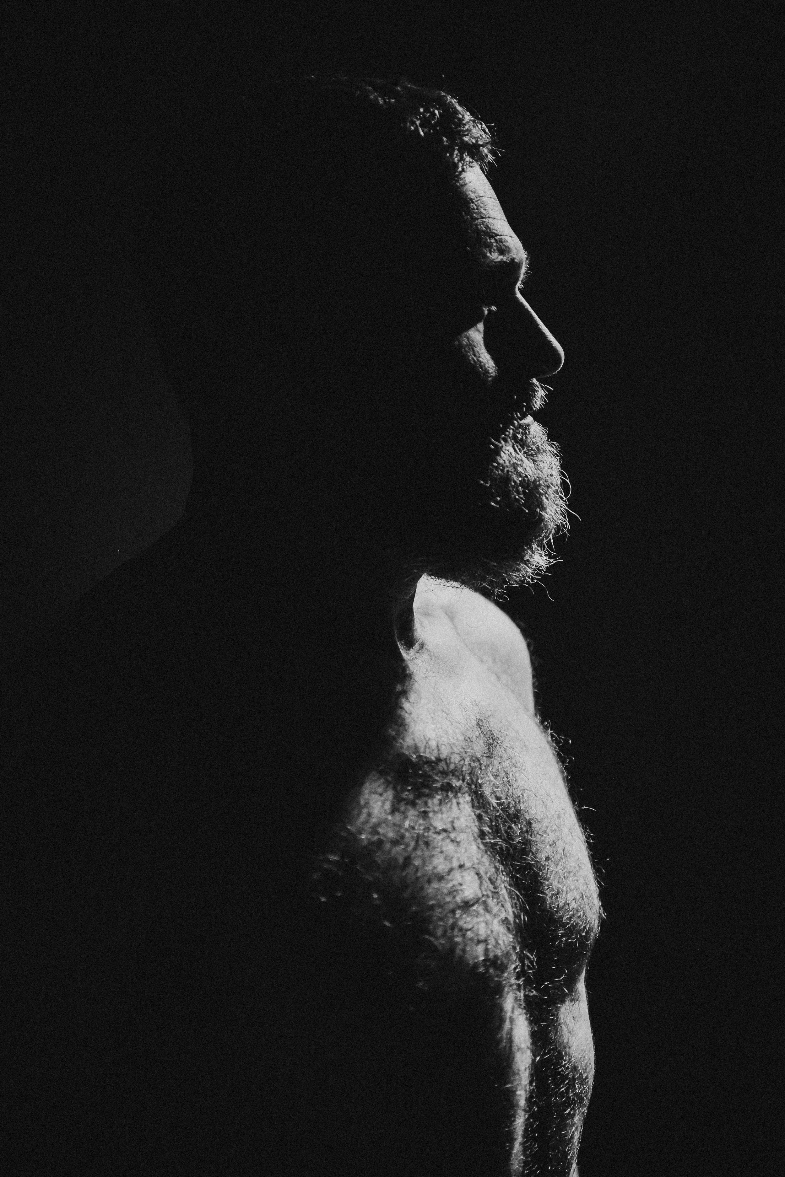 Side profile of a bearded man with minimal lighting highlighting his face and chest against a dark background.