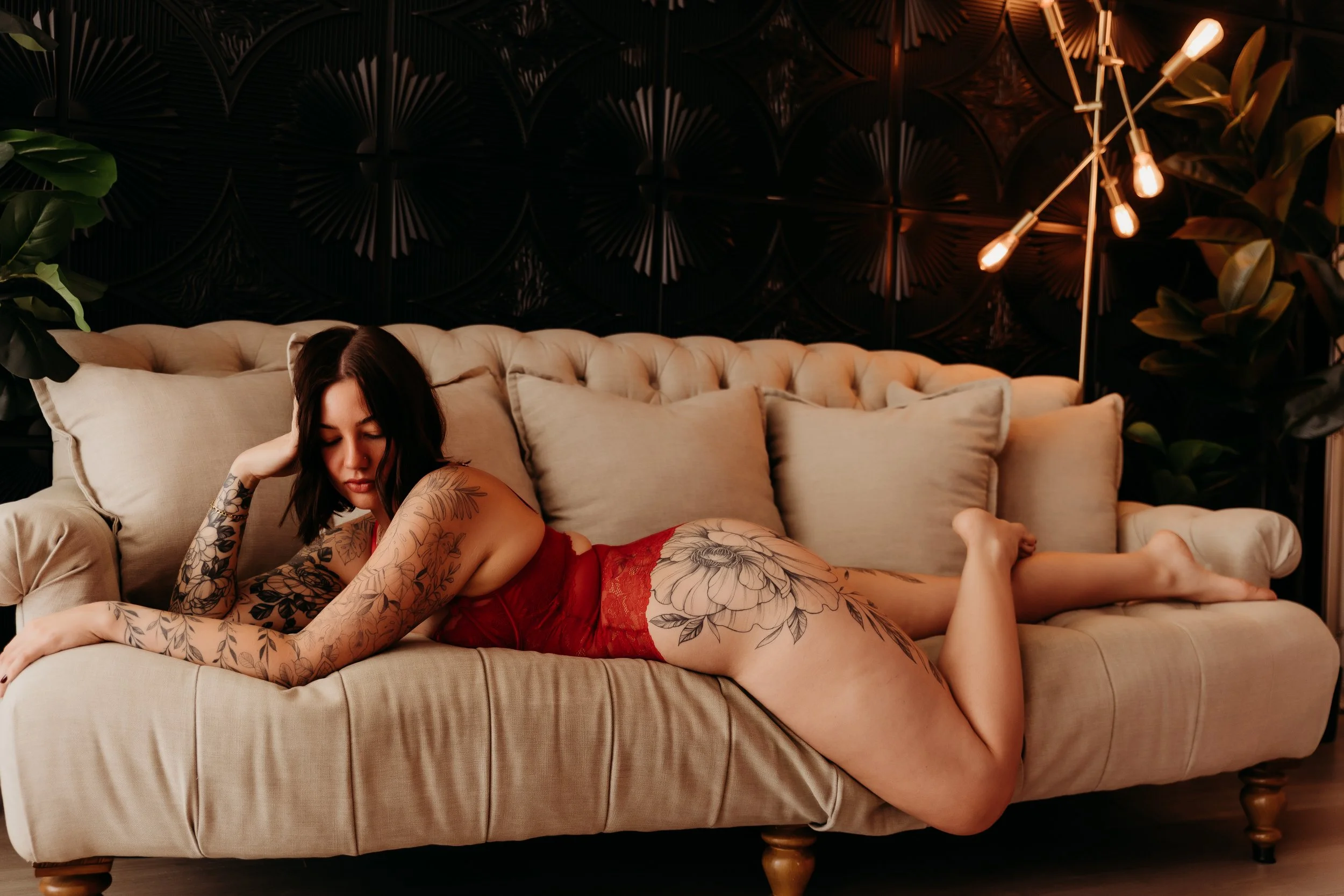 A woman with tattoos lounging on a beige couch in a dimly lit, stylish room with dark walls, green plants, and a modern floor lamp with exposed bulbs.