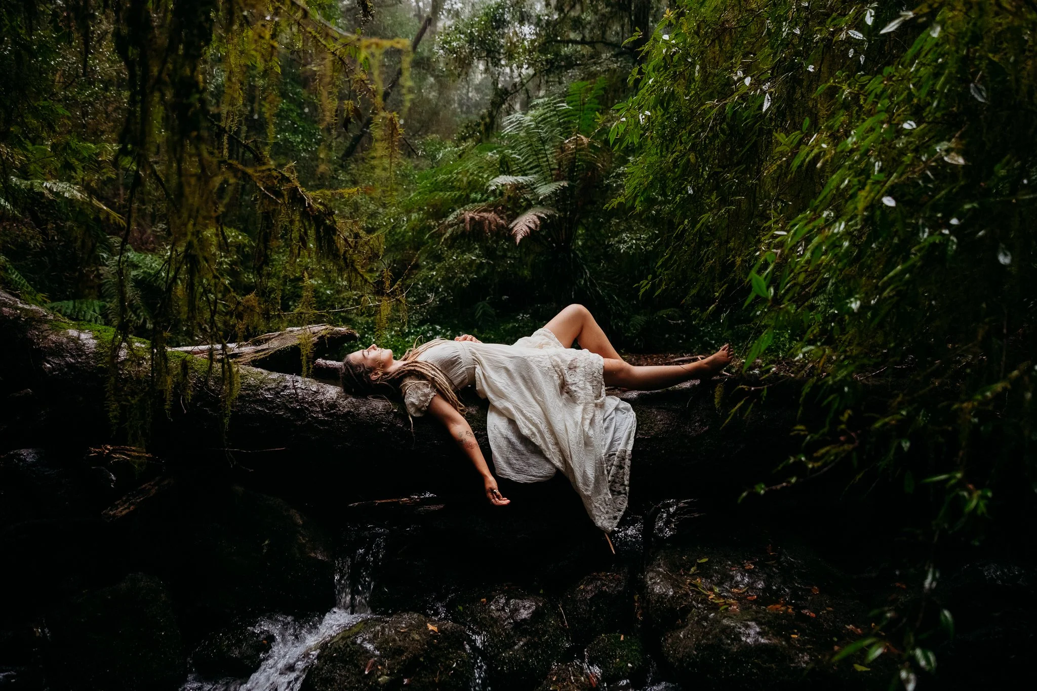 A woman in a white dress peacefully lying on a fallen tree trunk in a lush, green forest, with water flowing beneath.
