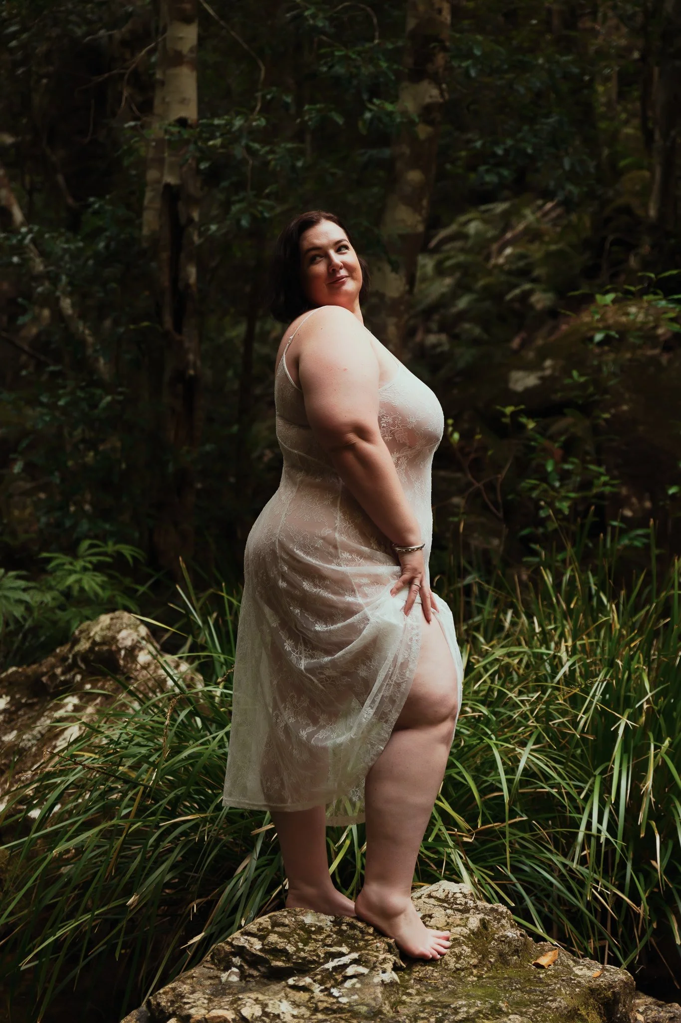 A woman with dark hair stands barefoot on a rock in a forested area, looking over her shoulder with a slight smile, wearing a and semi-transparent lace dress.