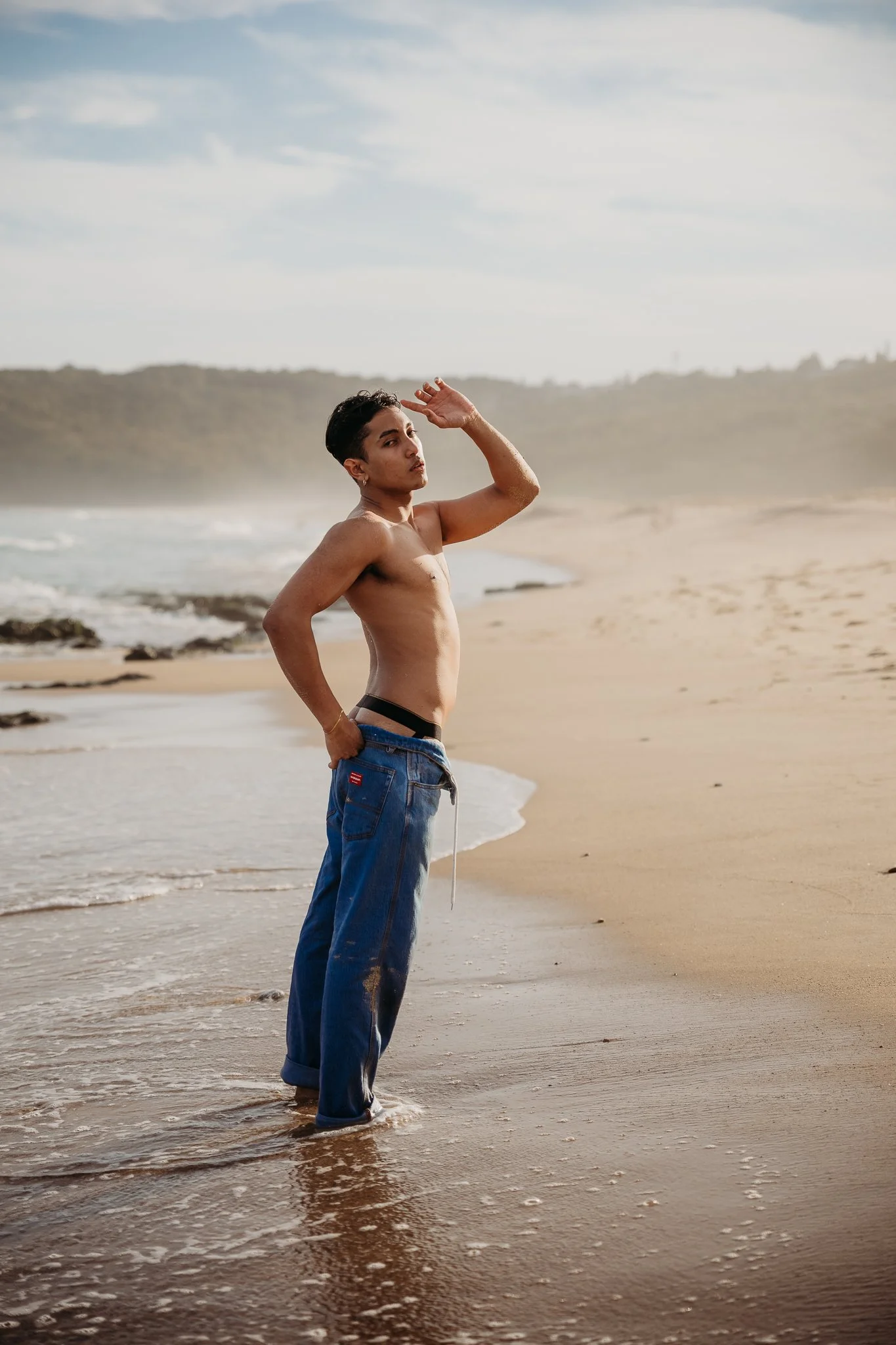 A shirtless young man standing at the edge of the ocean on a sandy beach, with one hand on his hip and the other shielding his eyes from the sun, wearing blue jeans.