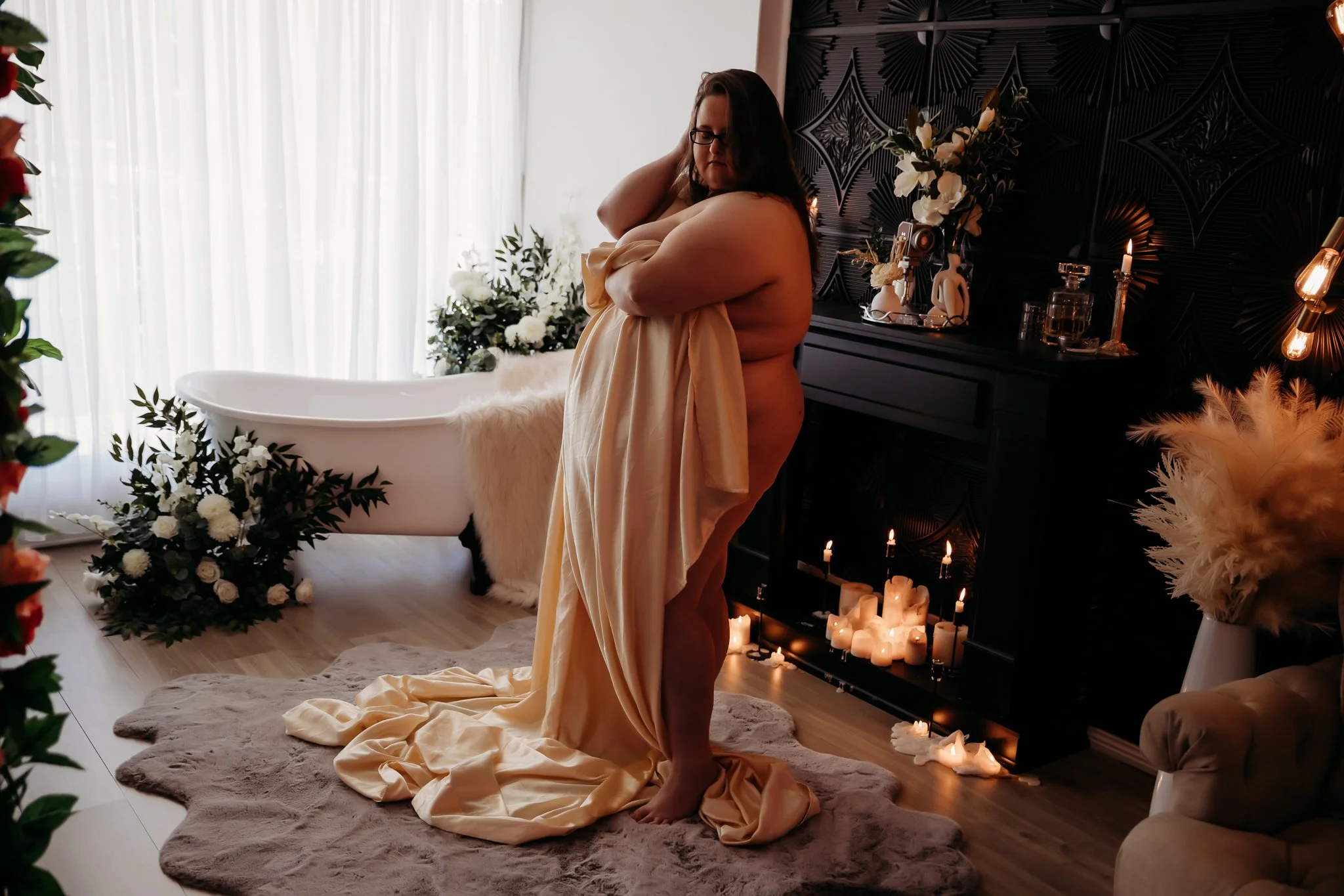 A woman standing on a plush rug in an elegantly decorated room, holding a piece of fabric, with a white bathtub decorated with flowers and candles in the background.