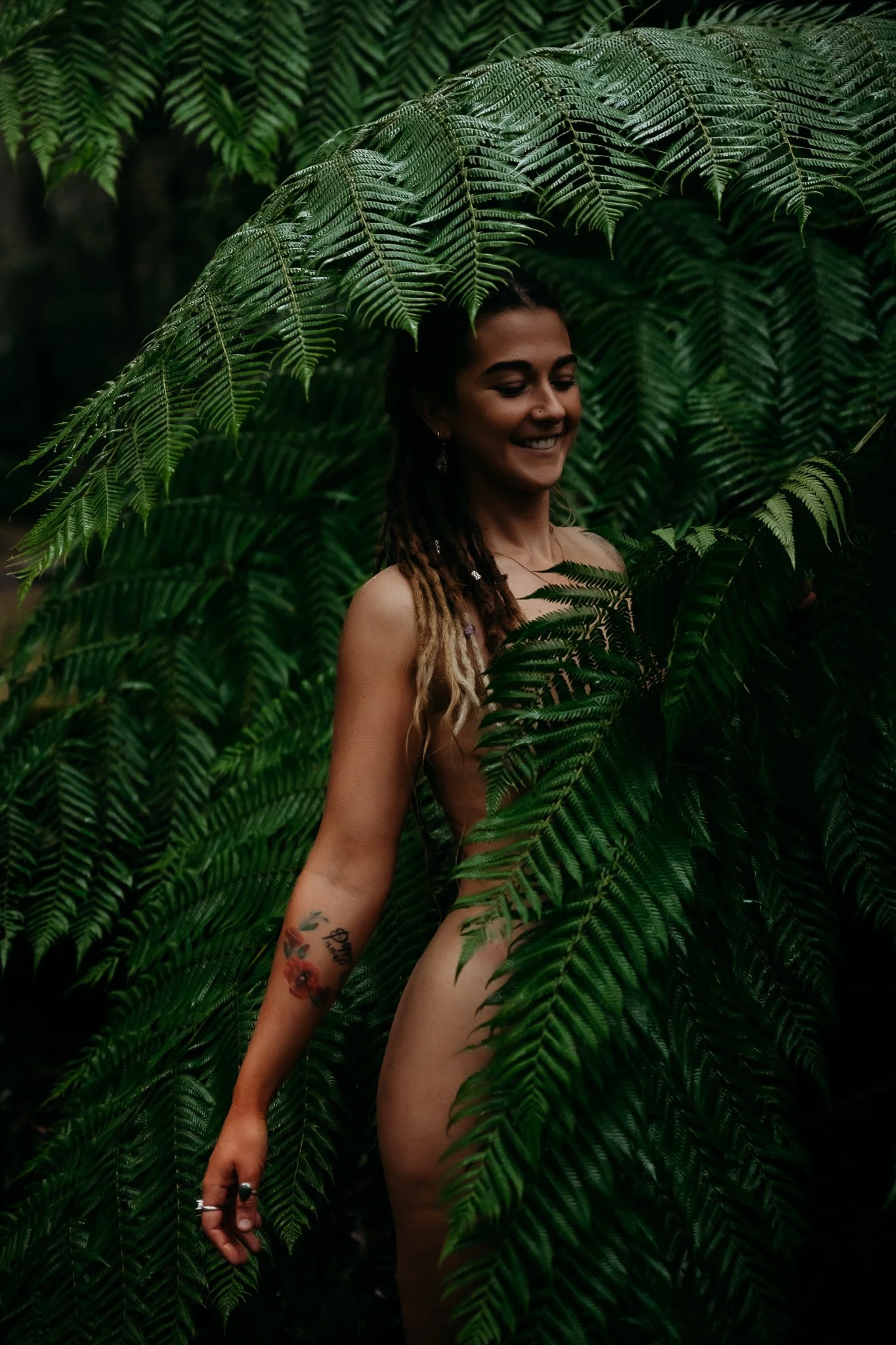 A woman with dreadlocks, tattoos, and jewelry stands behind lush green fern leaves, smiling with her eyes closed.