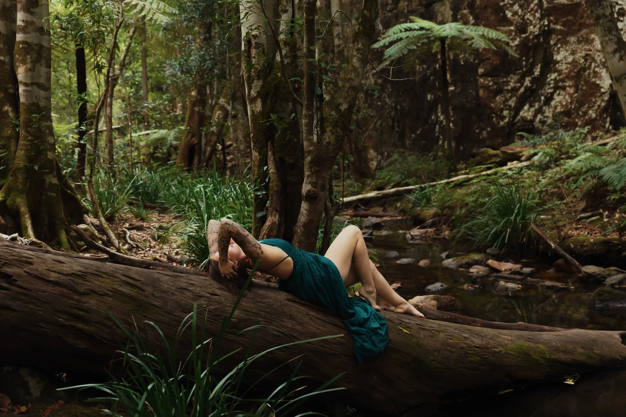 A woman laying on a fallen tree trunk in a lush forest with green foliage and a small stream.