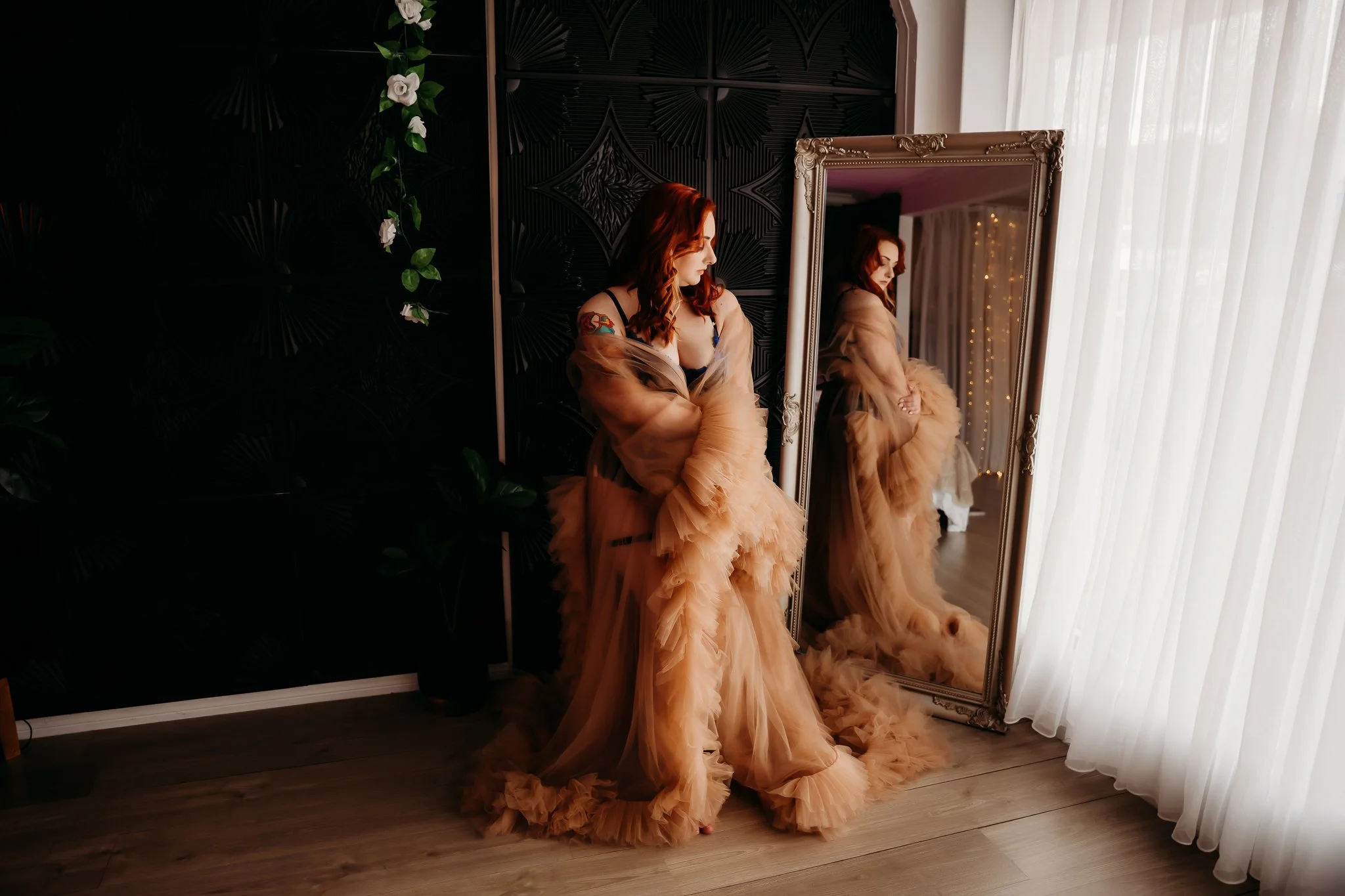 A woman with red hair wearing a beige ruffled gown stands in front of a tall mirror, adjusting her dress. The room has sheer white curtains on the right and a black textured wall with a plant on the left.