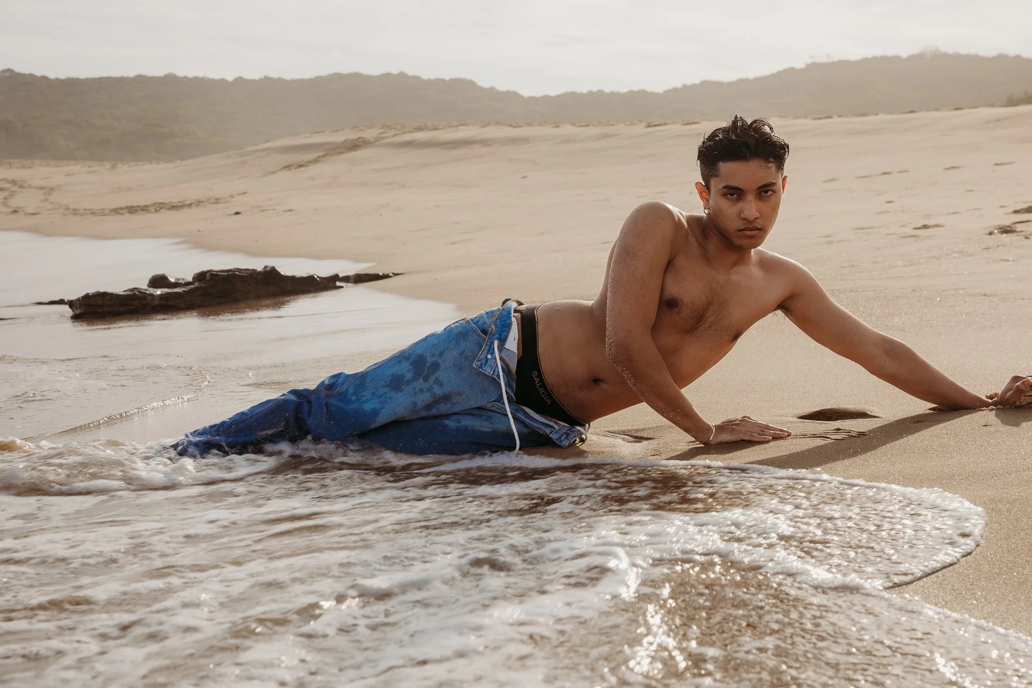 A shirtless young man in blue jeans lying on a sandy beach with waves at his side, looking directly at the camera.