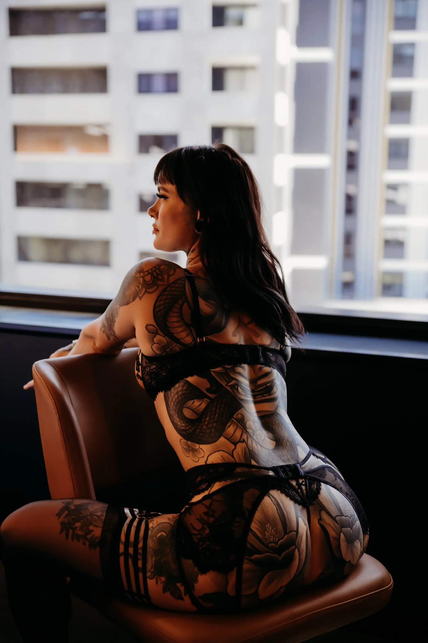 A woman with dark hair and tattoos sitting on a chair by a window, wearing black lace lingerie, with an urban cityscape outside.