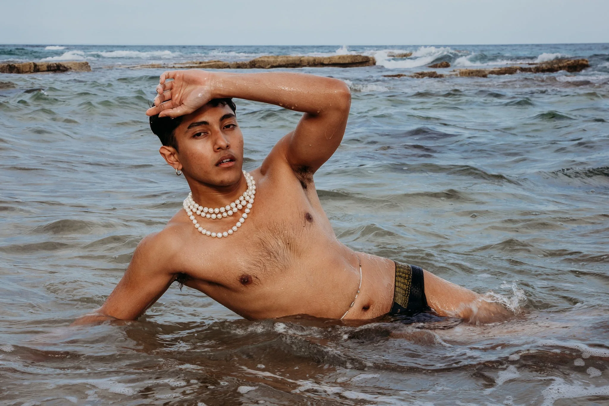 A shirtless man with pearl necklaces and earrings is lying in shallow ocean water near rocks, with one hand on his head and looking at the camera.