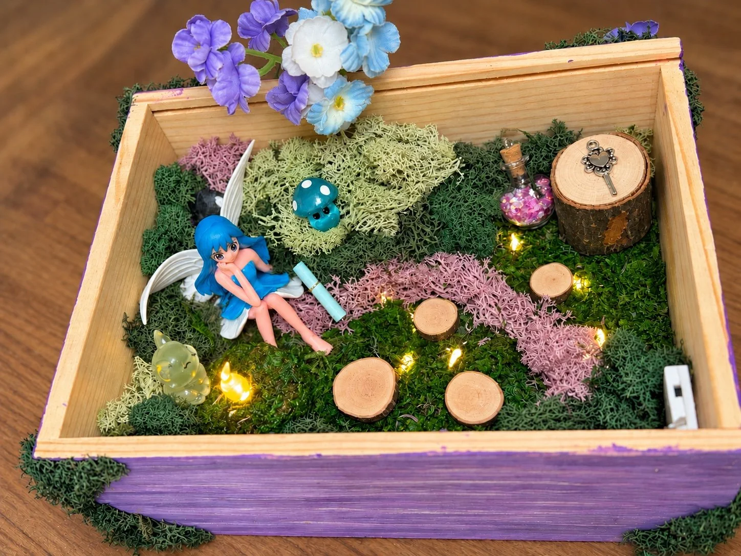 Finished TulleY Crafty fairy garden in a purple wooden box with moss, flowers, fairy lights, and a blue-haired fairy.