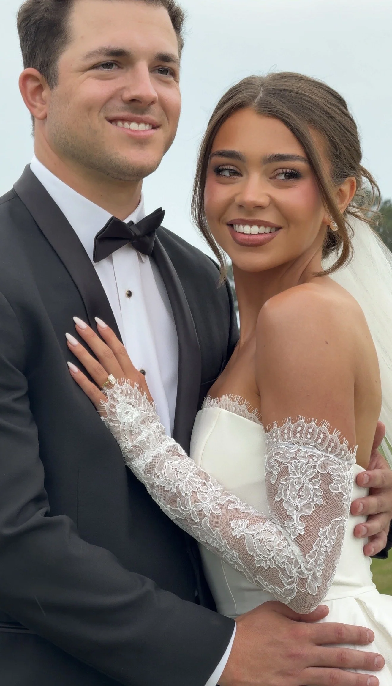 A newlywed couple smiling, with the groom in a black tuxedo and the bride in a white wedding dress with lace sleeves, outdoors on a cloudy day.