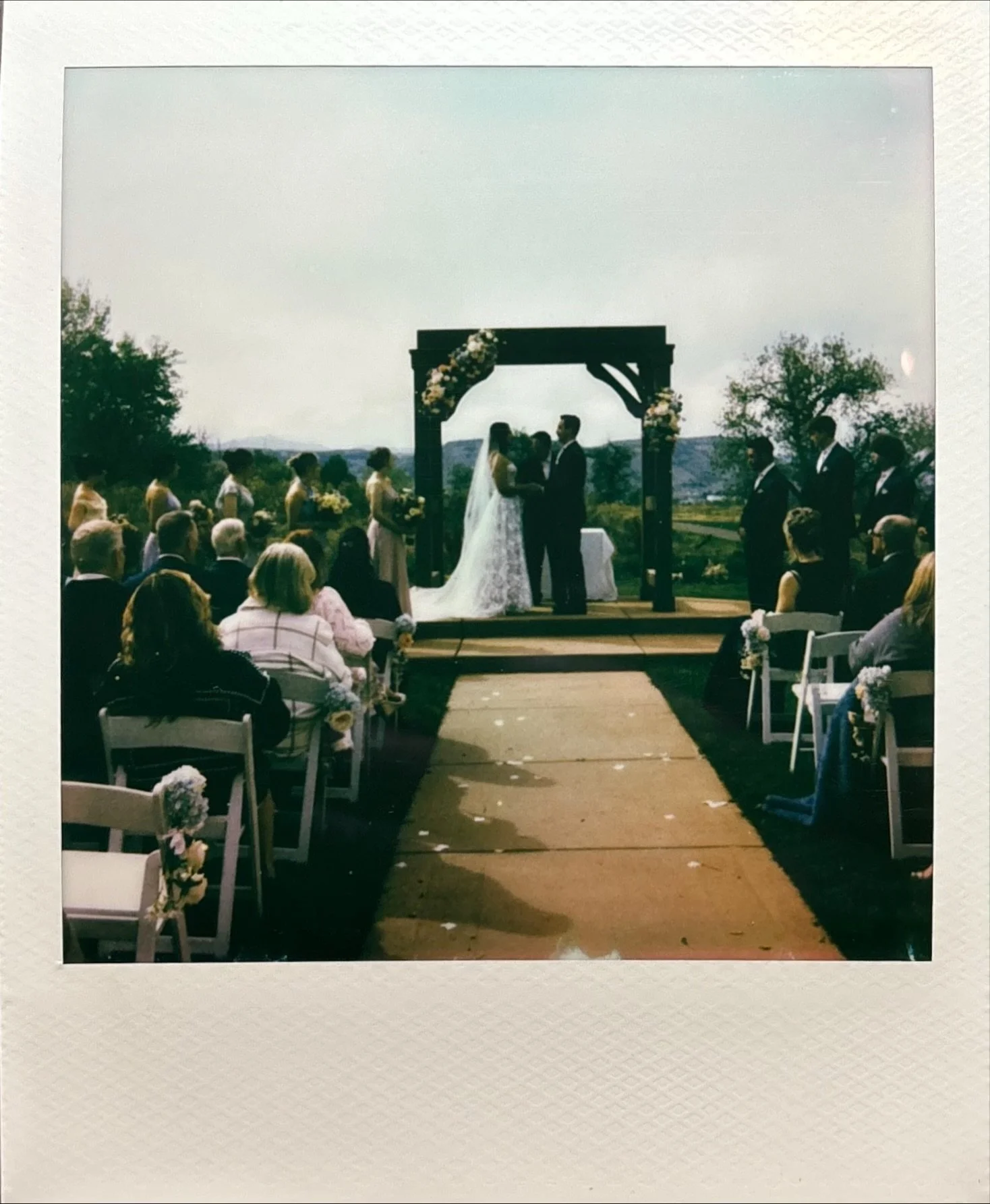 A wedding ceremony taking place outdoors under a wooden arch with floral decorations. The bride and groom are embracing and kissing in the center, with guests seated on either side of an aisle.
