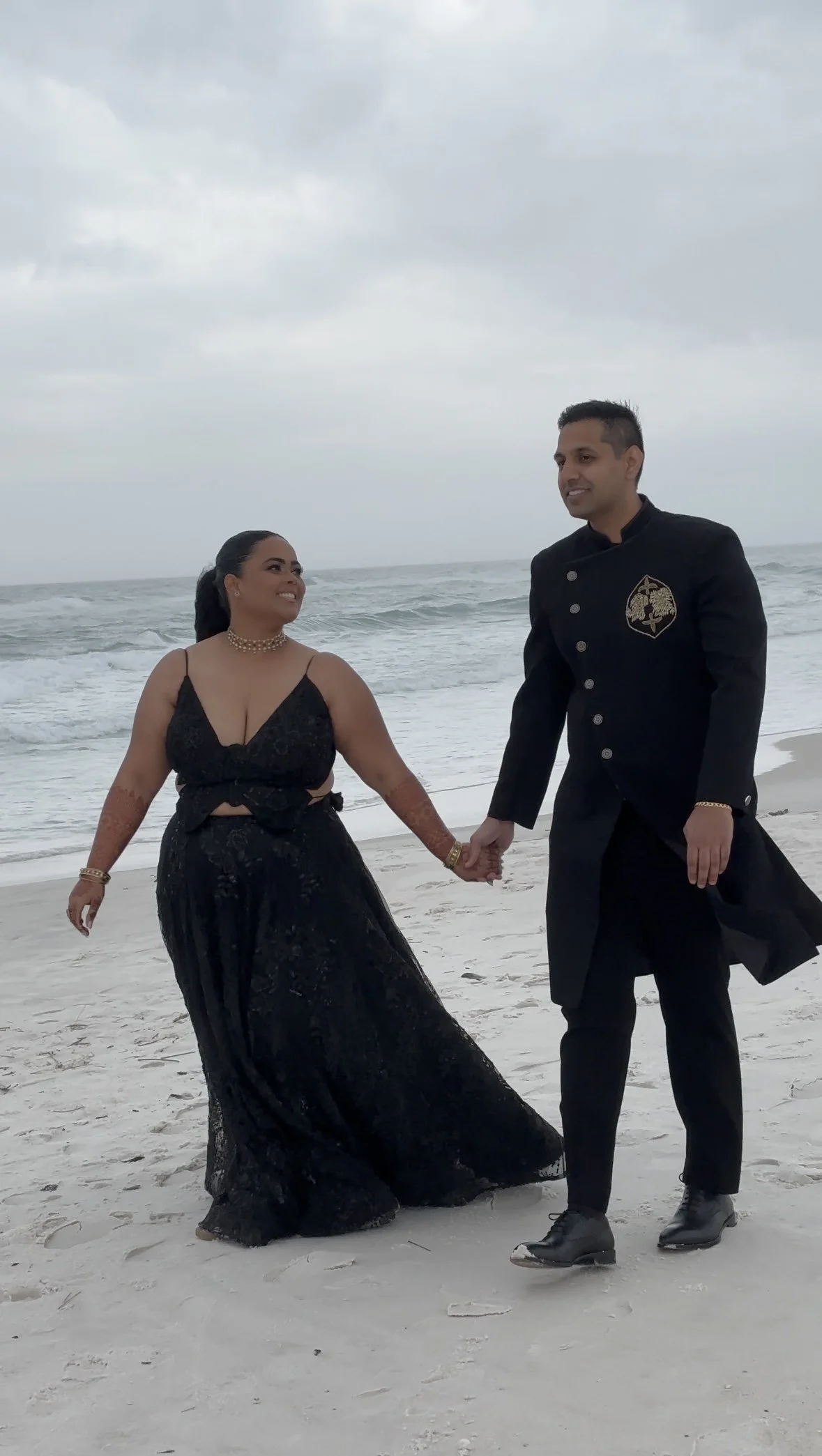 A couple holding hands walks on a beach with gray clouds overhead. The woman wears a black lace dress and jewelry, and the man wears a black uniform with a badge.