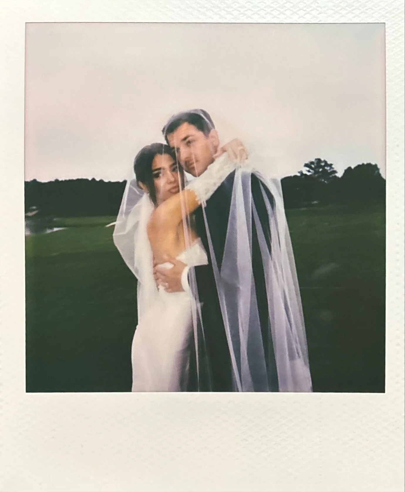 A bride and groom embracing outdoors during a wedding, with the bride in a white dress and the groom in a black suit, holding each other close.