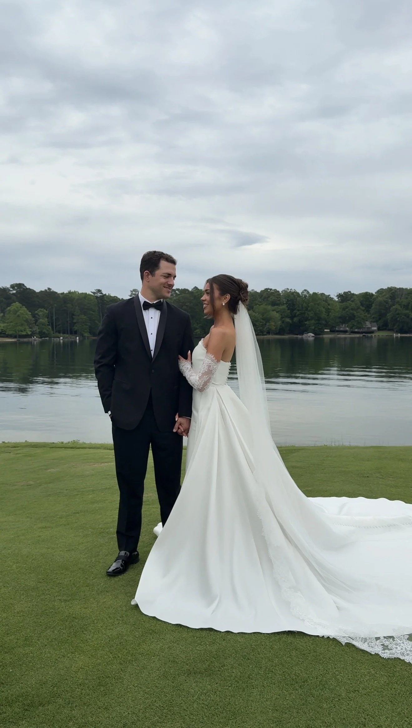 A bride and groom in wedding attire holding hands by a lake, smiling at each other, with cloudy sky and trees in the background.