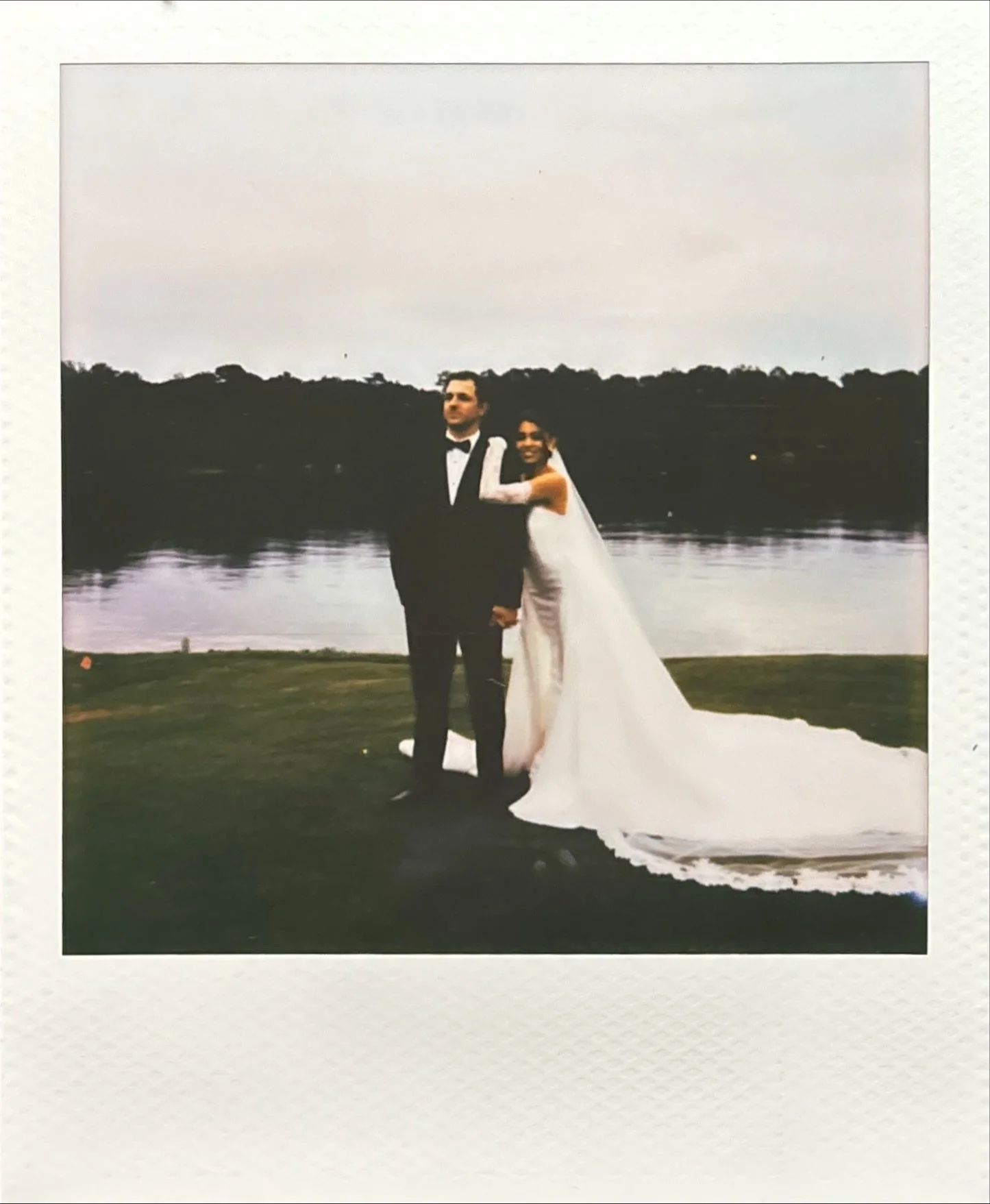 A wedding photo of a couple standing near a river, with trees in the background, the groom in a black tuxedo and the bride in a white wedding dress with a long train.