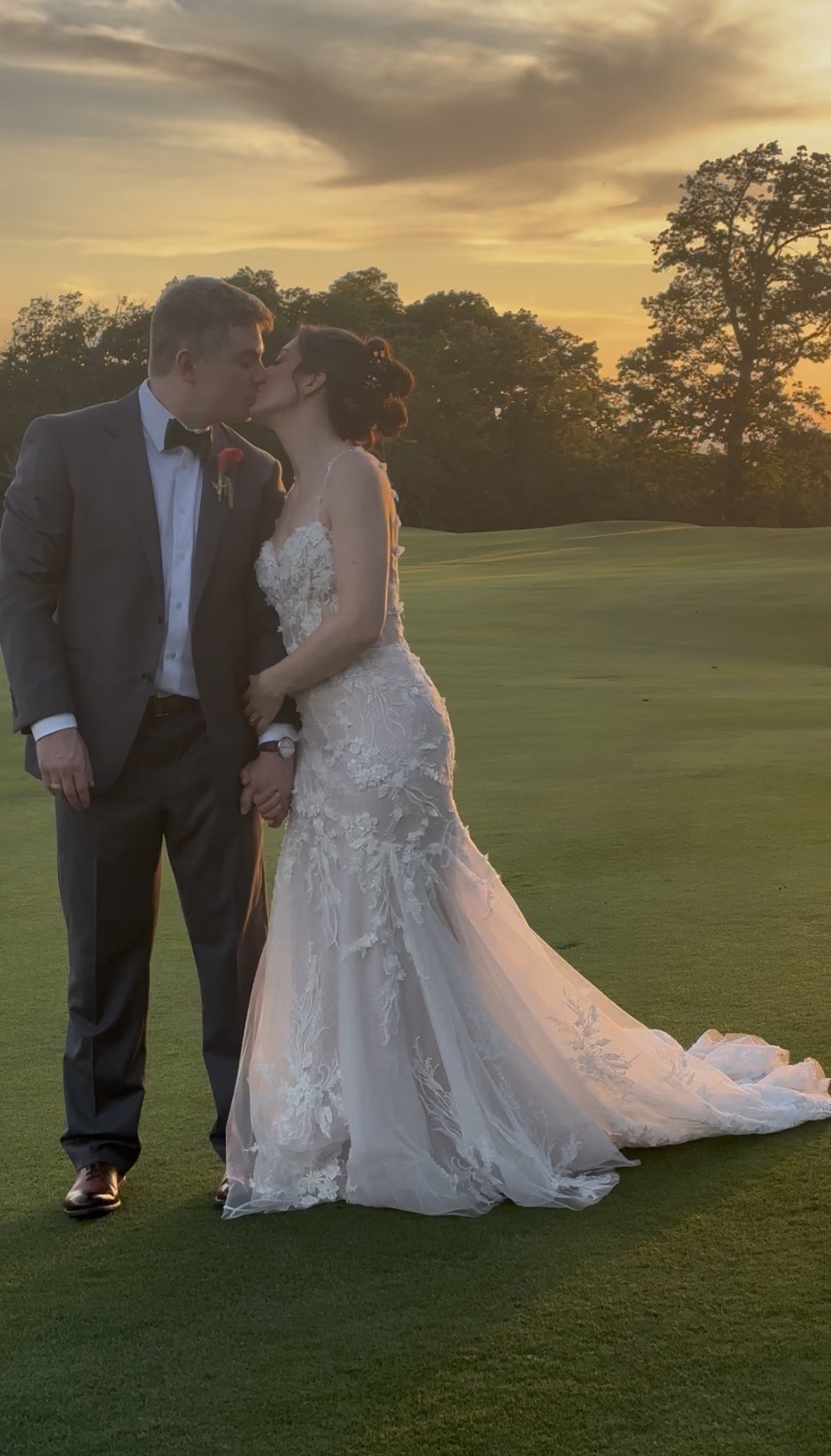 A bride and groom sharing a kiss on a golf course during sunset, with trees in the background and the bride wearing a lace wedding gown.