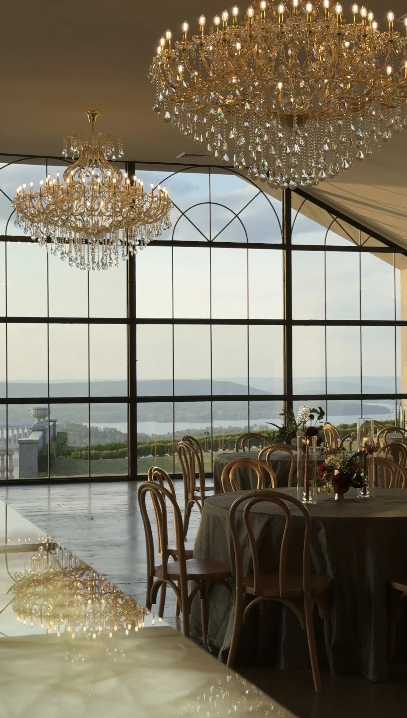 Elegant banquet hall with large crystal chandeliers, round tables with floral centerpieces, and large windows overlooking a scenic landscape with hills and water.