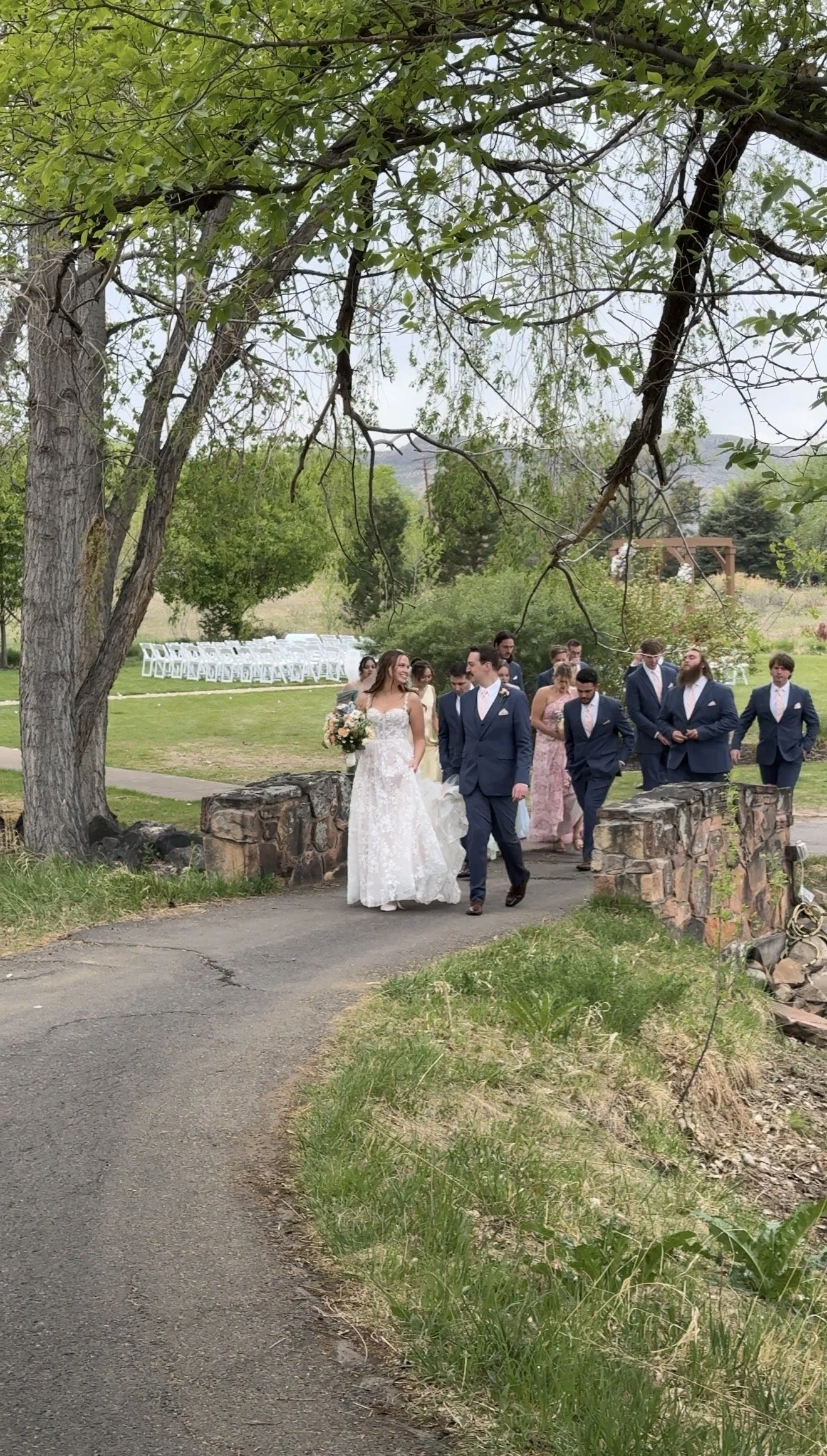 A bride and groom walking together on a path at an outdoor wedding ceremony, surrounded by guests and trees.
