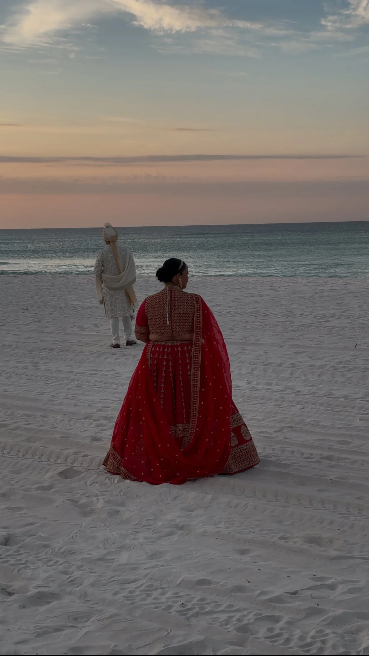 A woman in a red saree standing on a sandy beach looking towards the ocean, with a man in traditional white attire holding a staff, in the background during sunset.