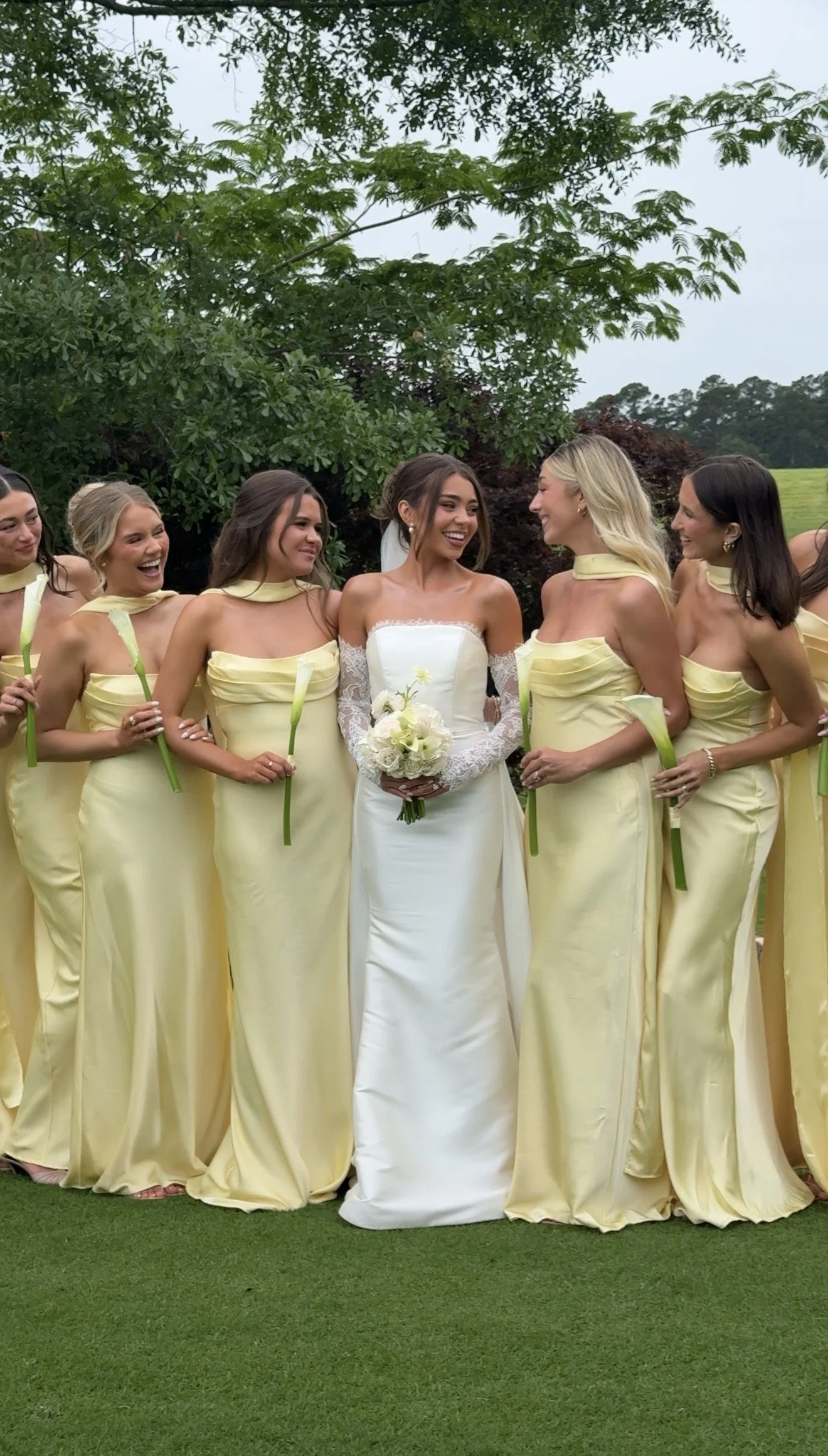 Group of women in yellow dresses, with a bride in a white gown holding a bouquet, standing outdoors on grass with trees in the background, celebrating a wedding.