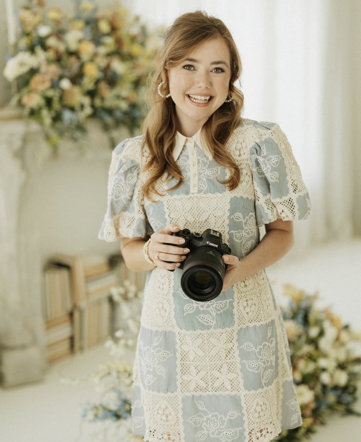 A young couple is having a romantic photoshoot outdoors near a lake with trees overhead, a woman in a white dress and tan boots, a man in a gray shirt and beige pants, and a woman in a pink dress taking photos.