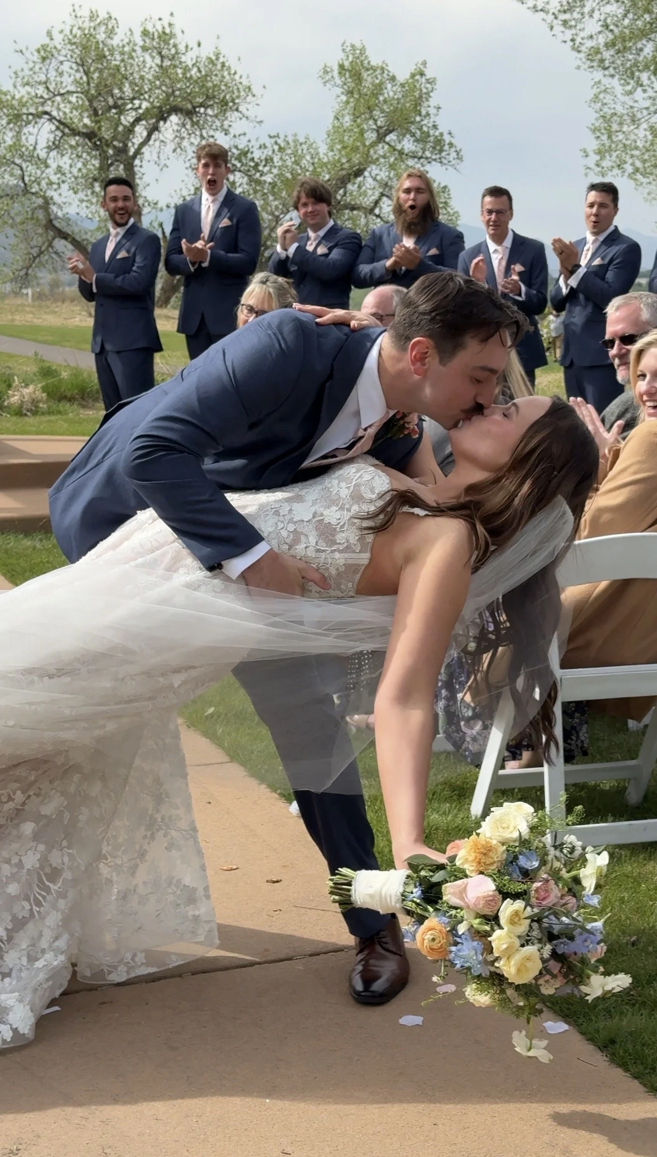 A bride and groom kiss at their outdoor wedding ceremony, with the bride lying on a chair and the groom leaning over her, while guests in suits applaud in the background. Flowers are scattered on the ground and a bouquet rests on the ground near the bride.