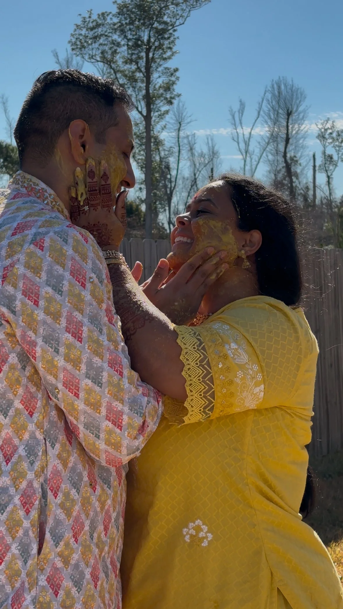 A couple celebrating a traditional Indian festival with turmeric paste on their faces and hands, outdoors with trees and a fence in the background.