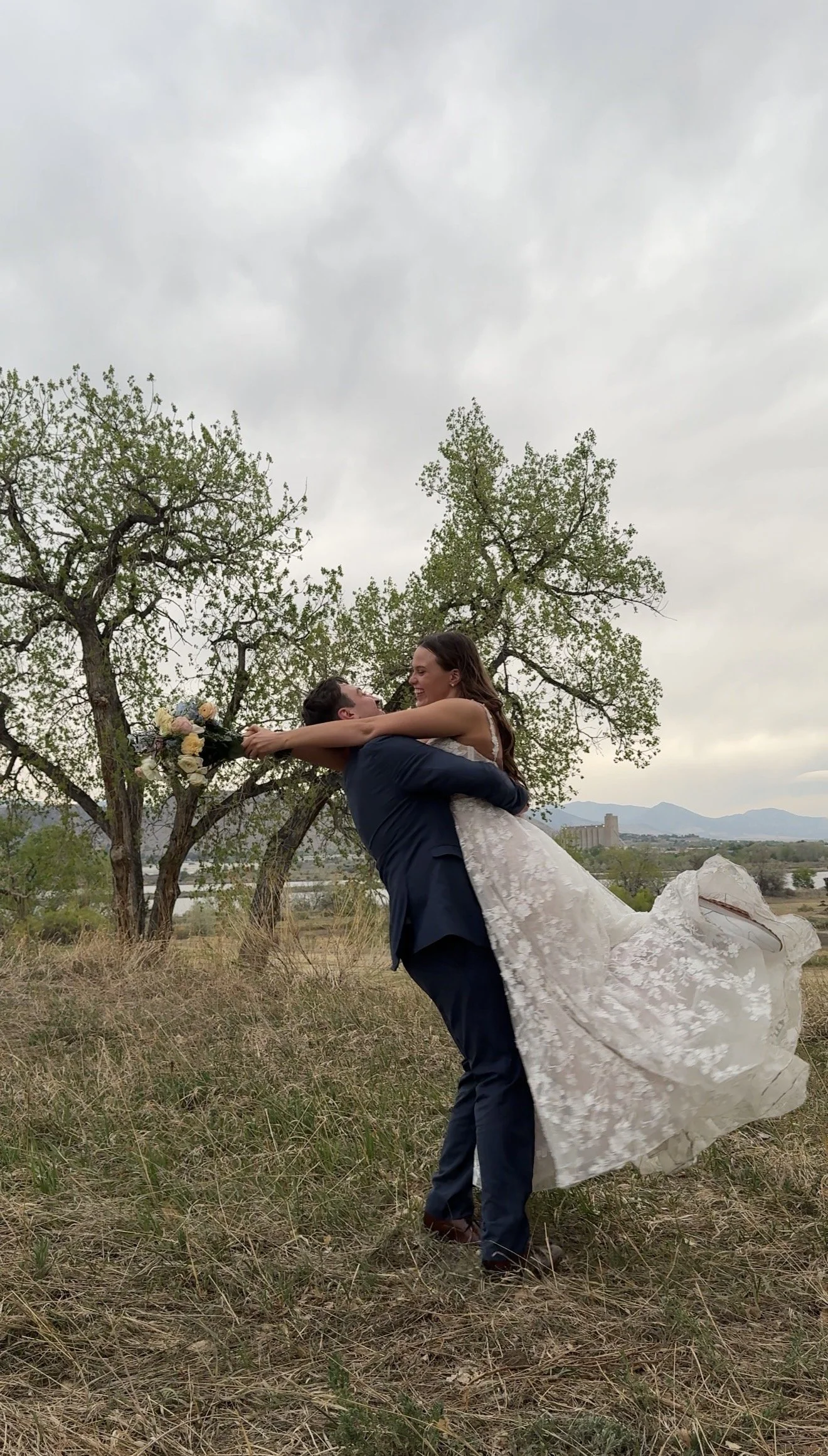 A bride and groom outdoors, with the groom lifting the bride in a joyful embrace. The bride is holding a bouquet, and both are smiling. The background features trees, distant mountains, and an overcast sky.