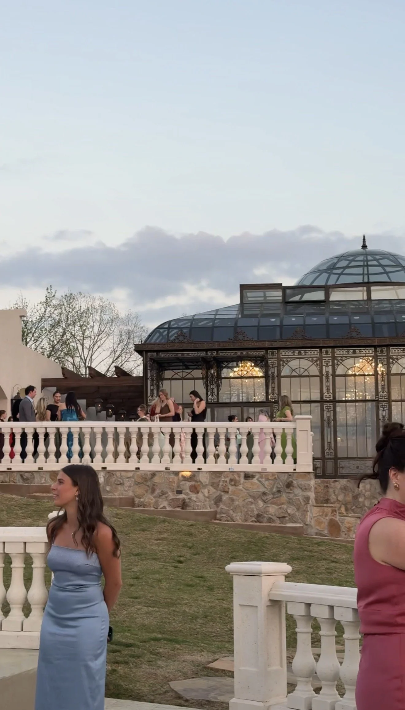 People gathered outdoors near a glass-roofed structure, with trees and clouds in the background, during sunset.