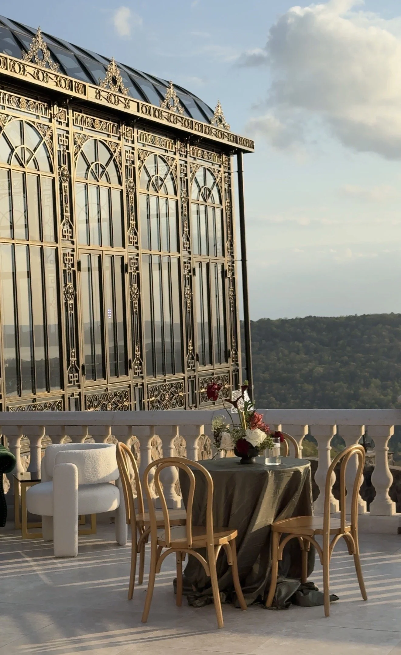 Elegant outdoor terrace with a decorative iron building facade, a table with a flower arrangement, chairs, and scenic hills in the background under a blue sky with clouds.