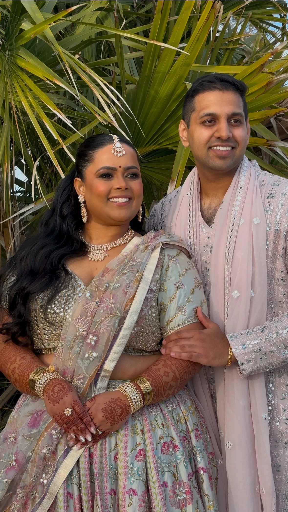A smiling couple dressed in traditional Indian wedding attire, standing outdoors in front of palm leaves.