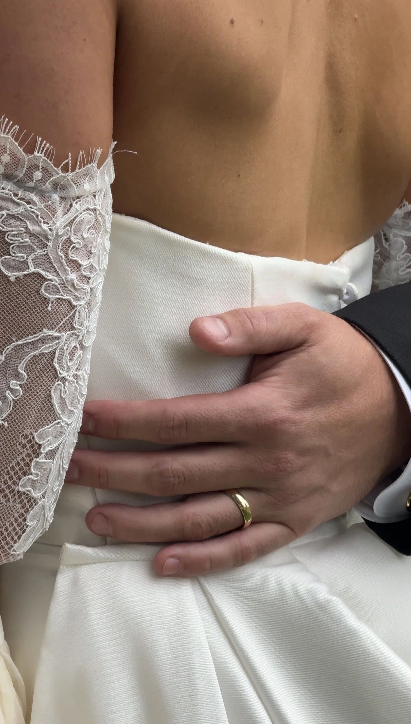Close-up of a couple's embrace during their wedding, showing the bride's lace sleeve and the groom's hand with a wedding band on his finger, resting on the bride's waist.