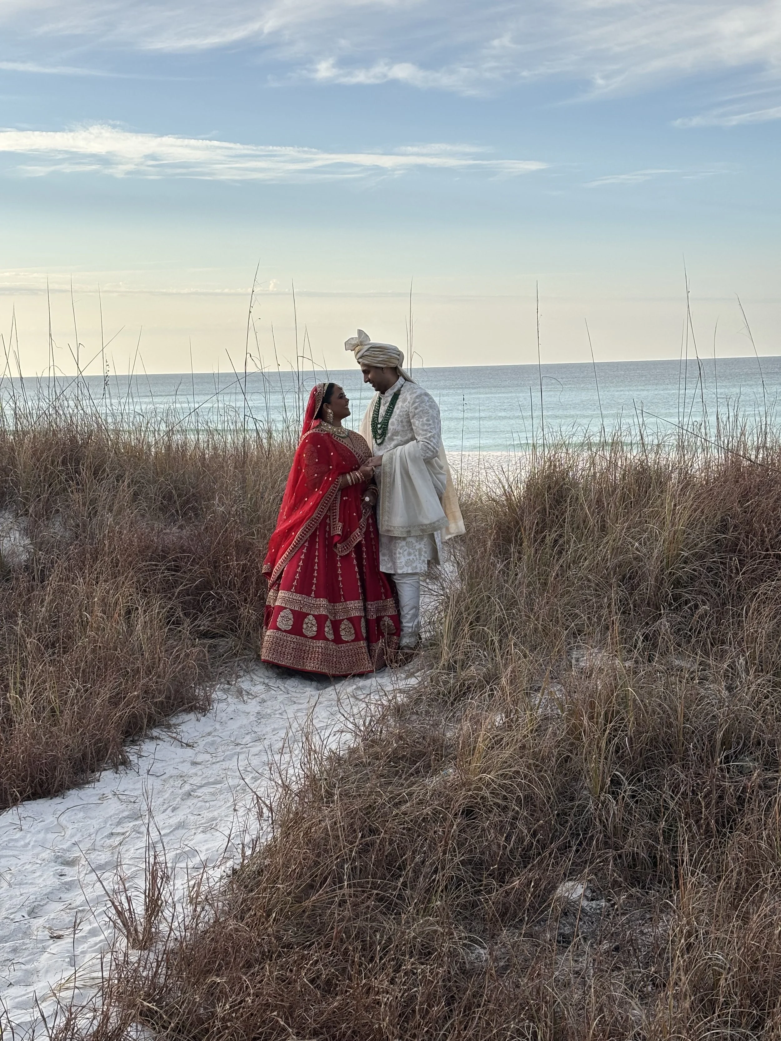 A bride and groom in traditional Indian wedding attire standing on a sandy path between tall beach grass, near the ocean with a partly cloudy sky.
