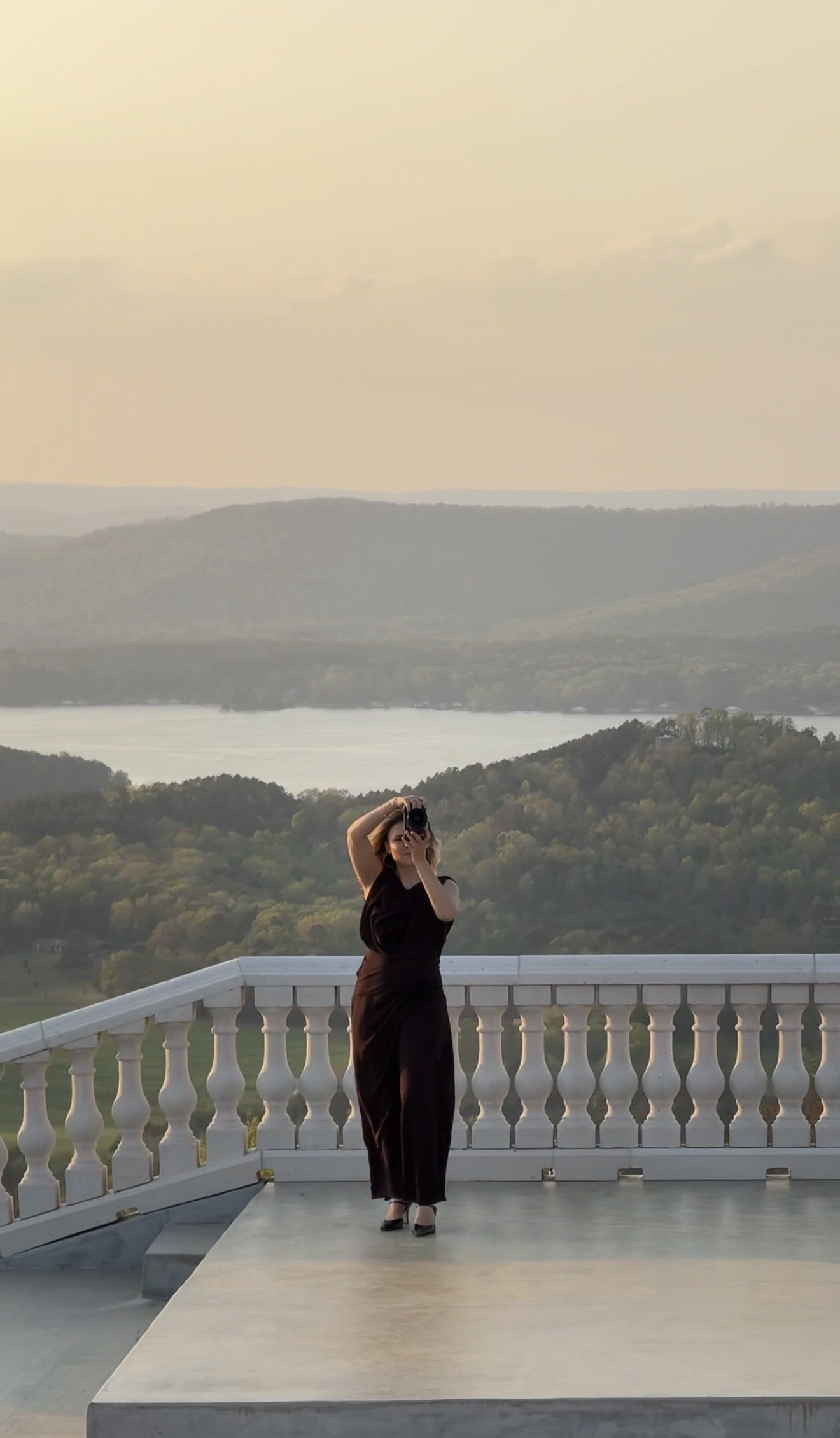 A woman in black dress and high heels standing on a white balcony with a scenic view of hills, water, and a cloudy sky in the background, taking a photo with a camera.