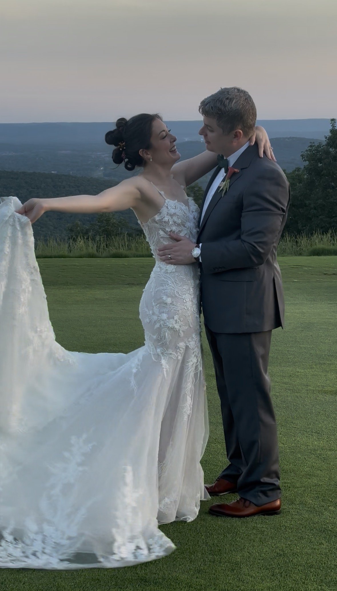 A bride and groom dancing outdoors on a grassy area with mountains in the background during sunset or dusk.