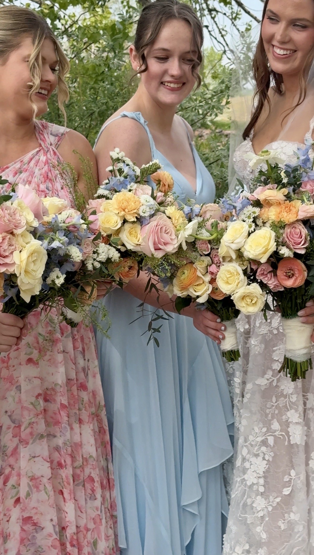 Three women standing outdoors in floral dresses, holding bouquets of mixed flowers, smiling and looking at each other.