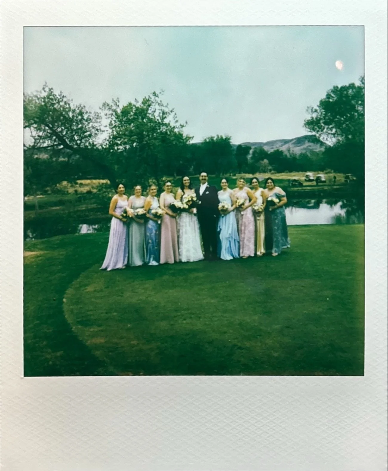 A group of ten people, including one man in a black tuxedo and nine women in colorful dresses holding bouquets, standing on lawn near a pond with trees and mountains in the background during daytime.