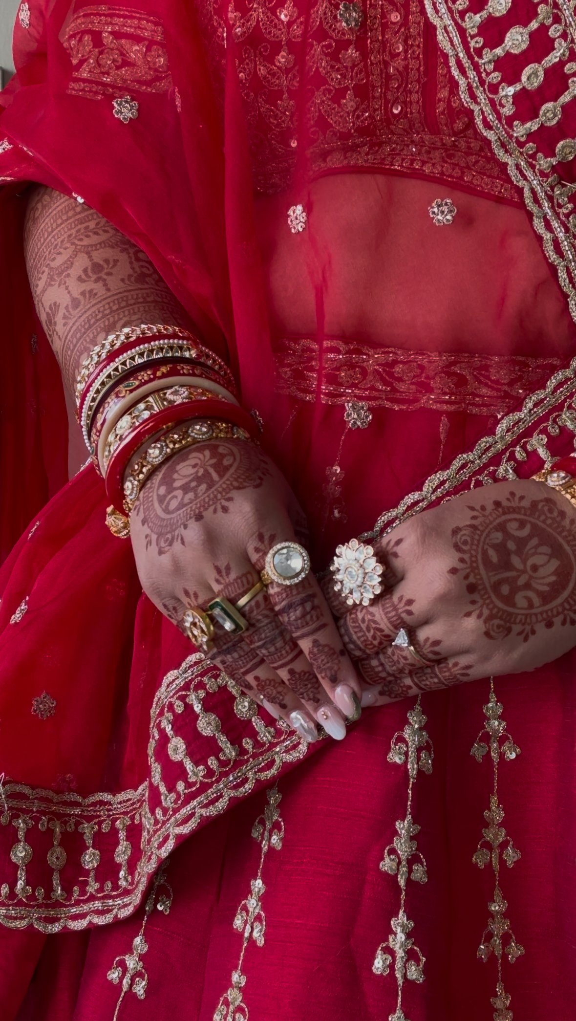Close-up of a woman in a red traditional Indian outfit with gold embroidery, wearing multiple gold and red bangles, rings, and henna designs on her hands and arms.