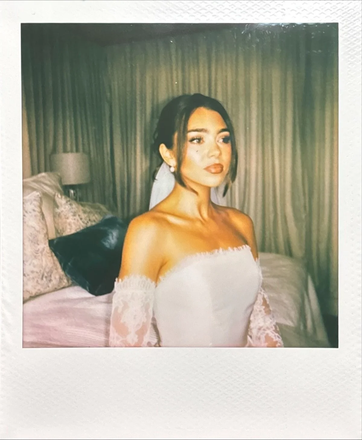 A woman with dark hair and makeup sitting on a bed in a hotel room, dressed in a white off-shoulder wedding gown with lace sleeves.