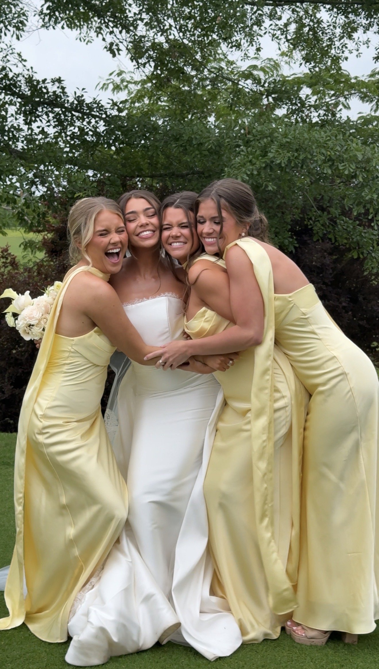 Five women, one in a wedding dress and four in yellow dresses, are hugging and smiling outdoors with trees in the background.