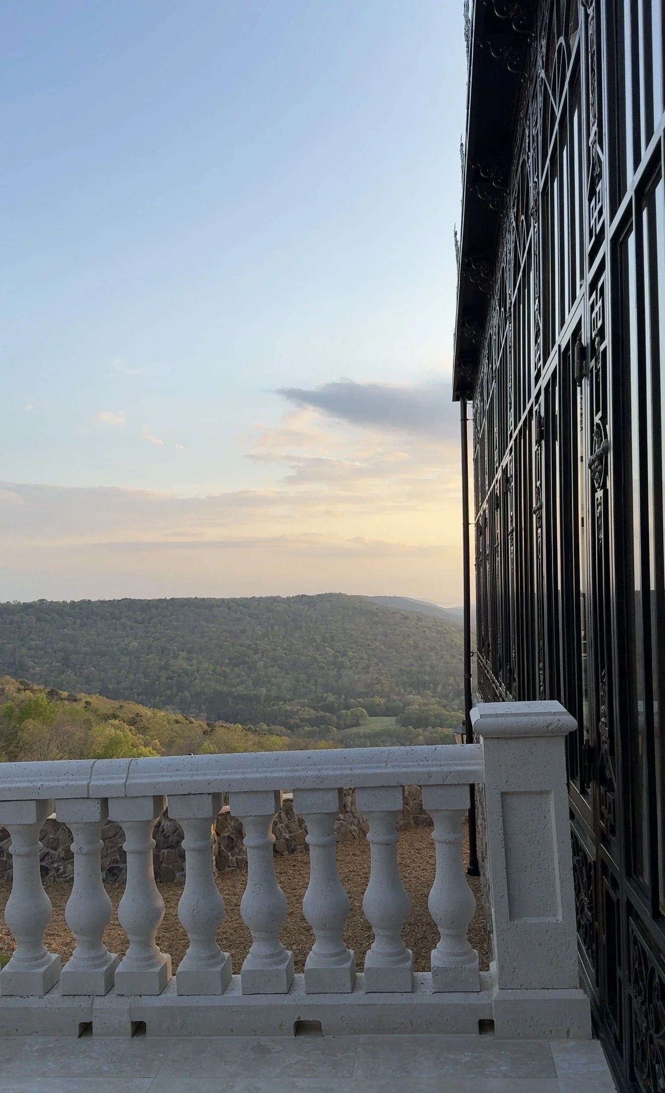Balcony with white balustrade overlooking green hills and a partly cloudy sky at sunset, with a black wrought iron railing on the right side.
