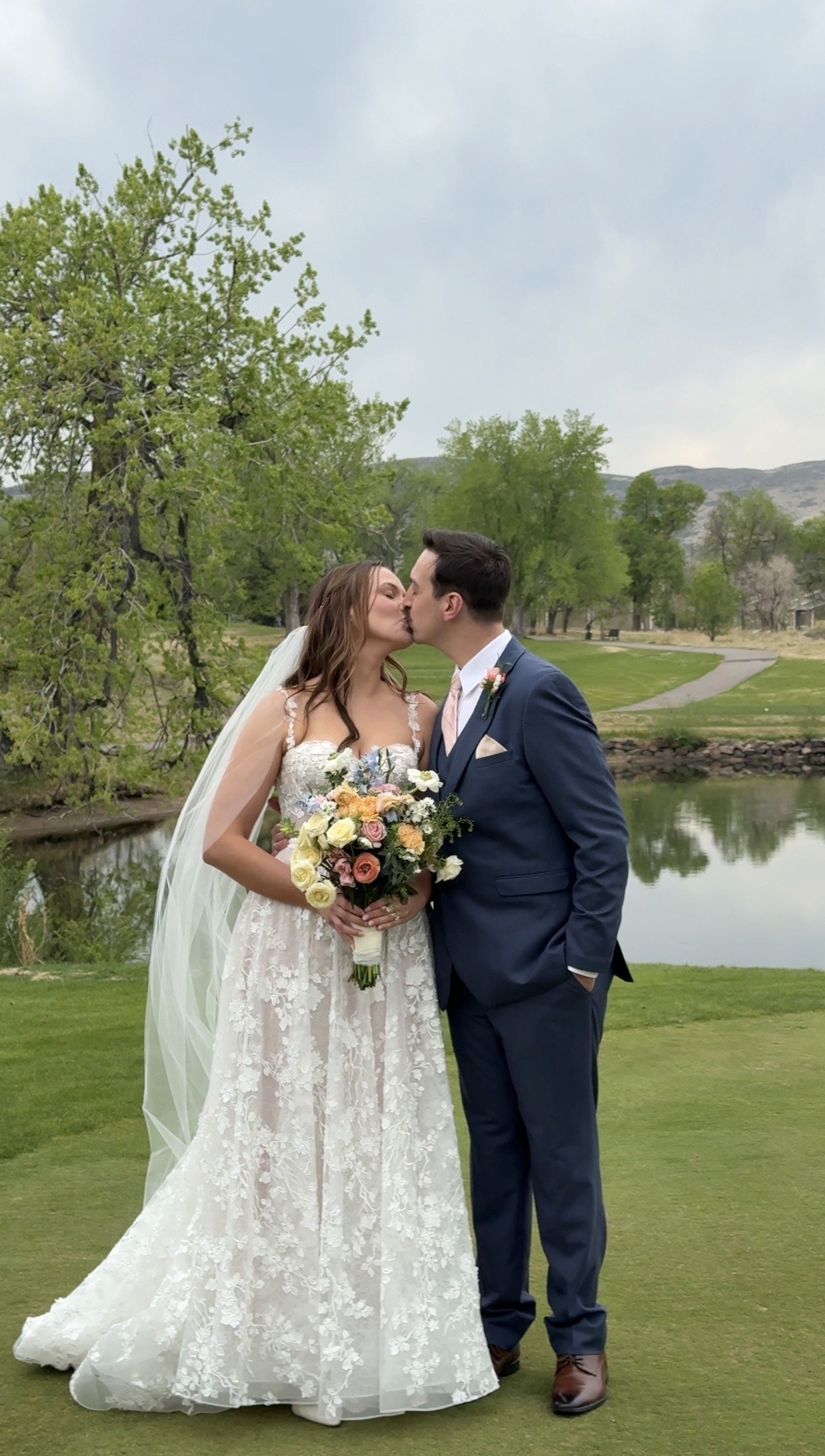 A bride and groom kissing outdoors on a grassy area near a pond, surrounded by green trees with mountains in the background.