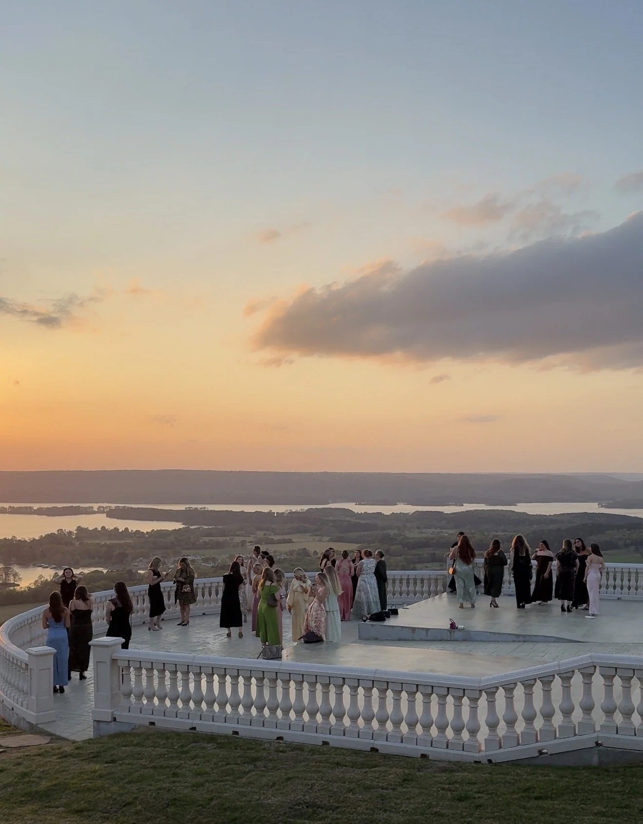People gathered on a white circular balcony overlooking a scenic landscape at sunset, with a river, fields, and a colorful sky with clouds.