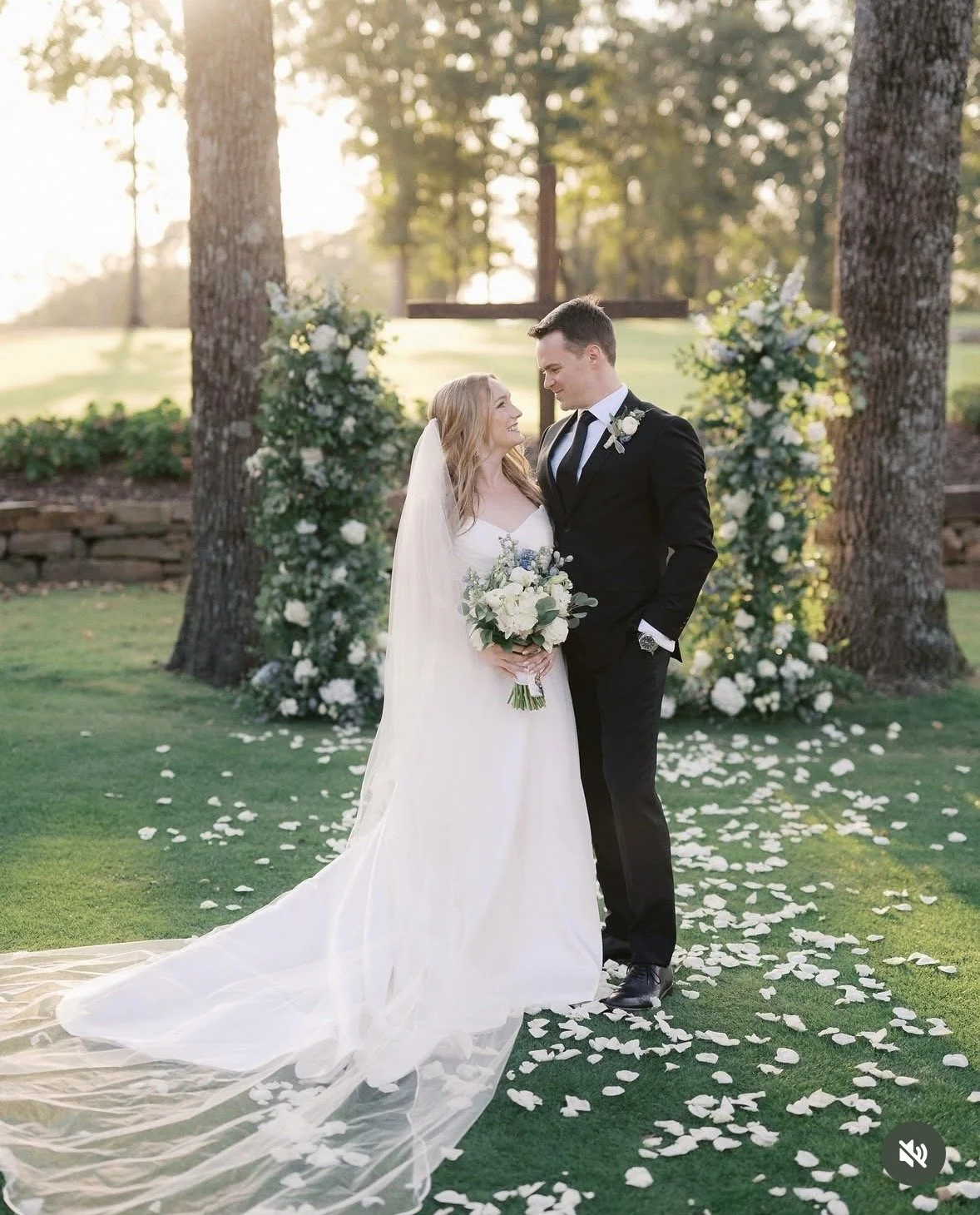 Bride in a strapless white wedding gown holding a bouquet of white roses and greenery, standing next to a groom in a black tuxedo with a white boutonniere, outside a stone building with arched windows and green bushes.