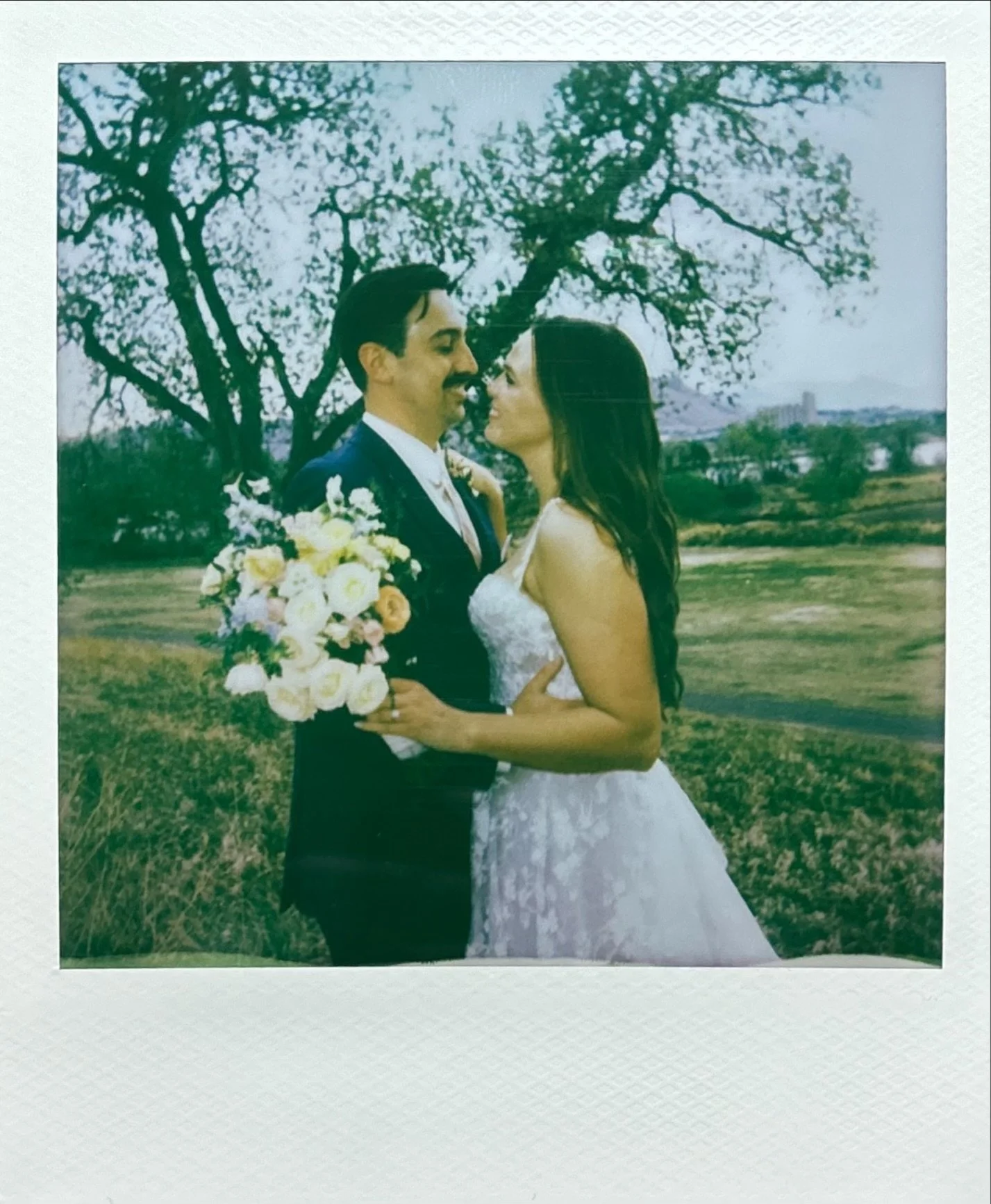 A couple on their wedding day standing outdoors by a tree, facing each other and smiling, with the groom holding a bouquet of flowers.