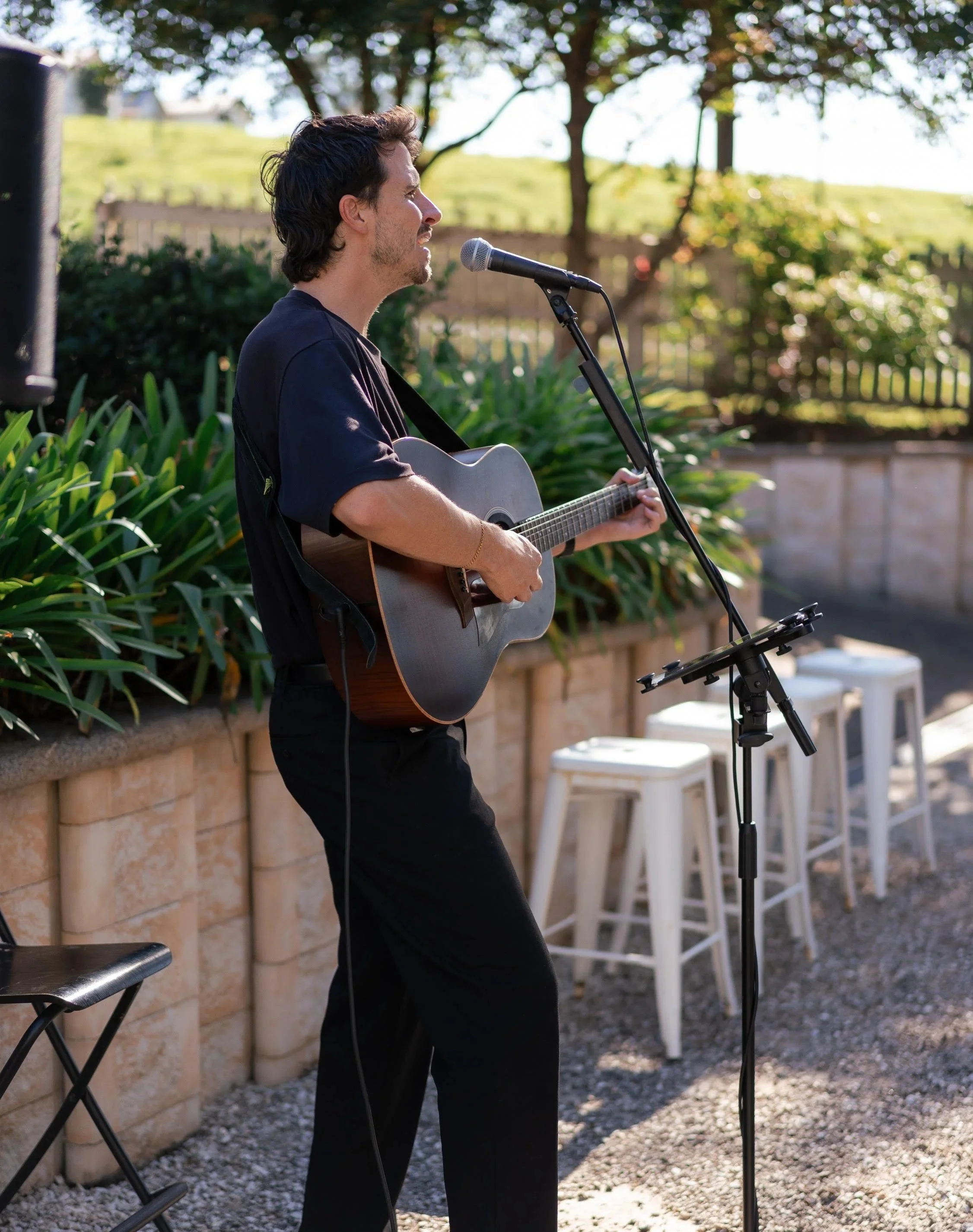 A man with dark curly hair, wearing a black t-shirt, playing an acoustic guitar and singing into a microphone outdoors during daytime with trees and a fence in the background.