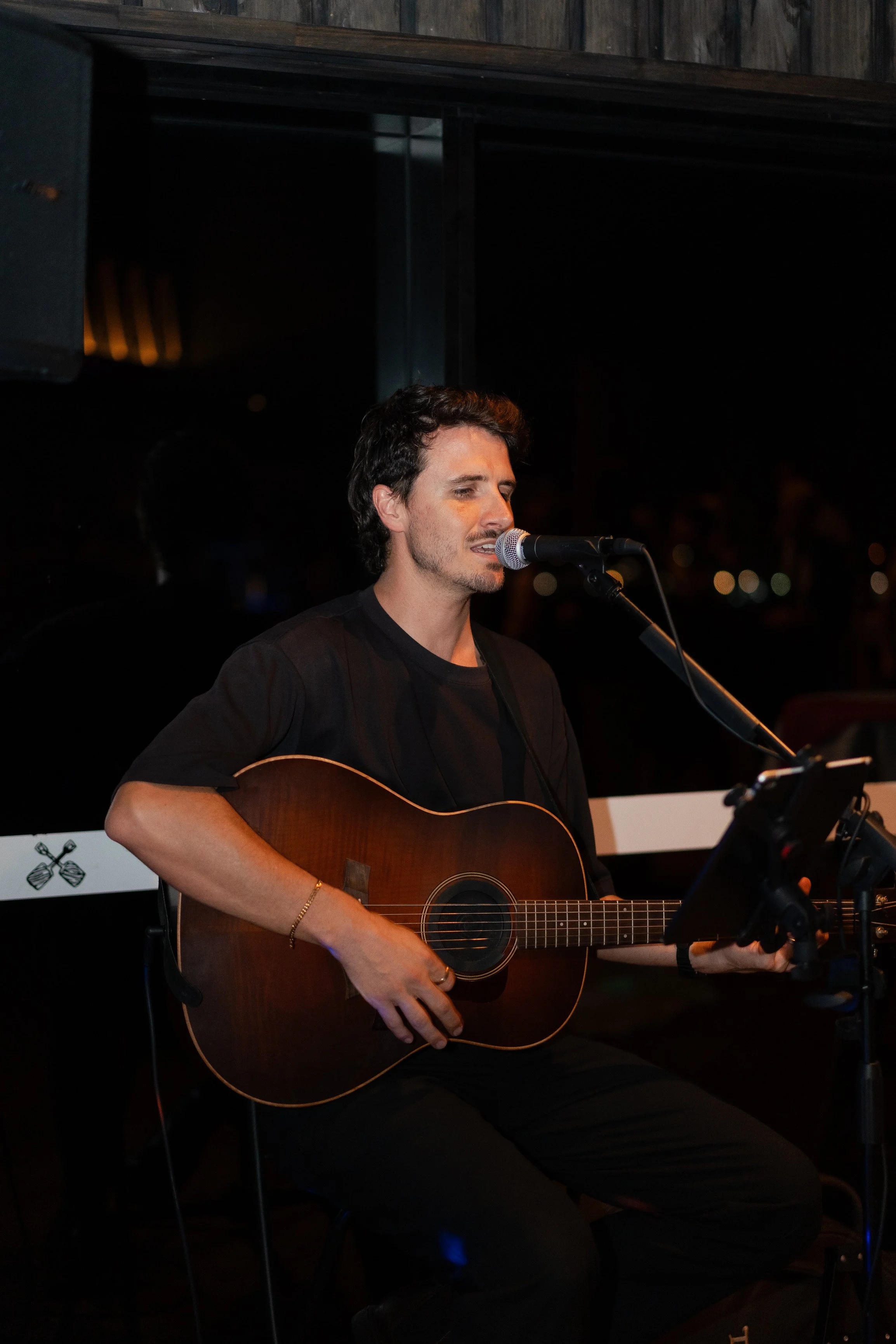 A man with dark hair and light skin is singing into a microphone while playing an acoustic guitar at night. He is wearing a black shirt and is seated in front of a keyboard, with a dark background and city lights visible in the distance.