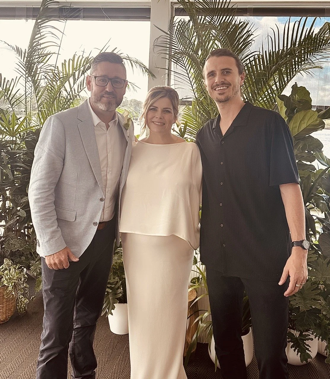 Three people smiling in front of potted plants indoors, dressed semi-formally, with a window in the background.