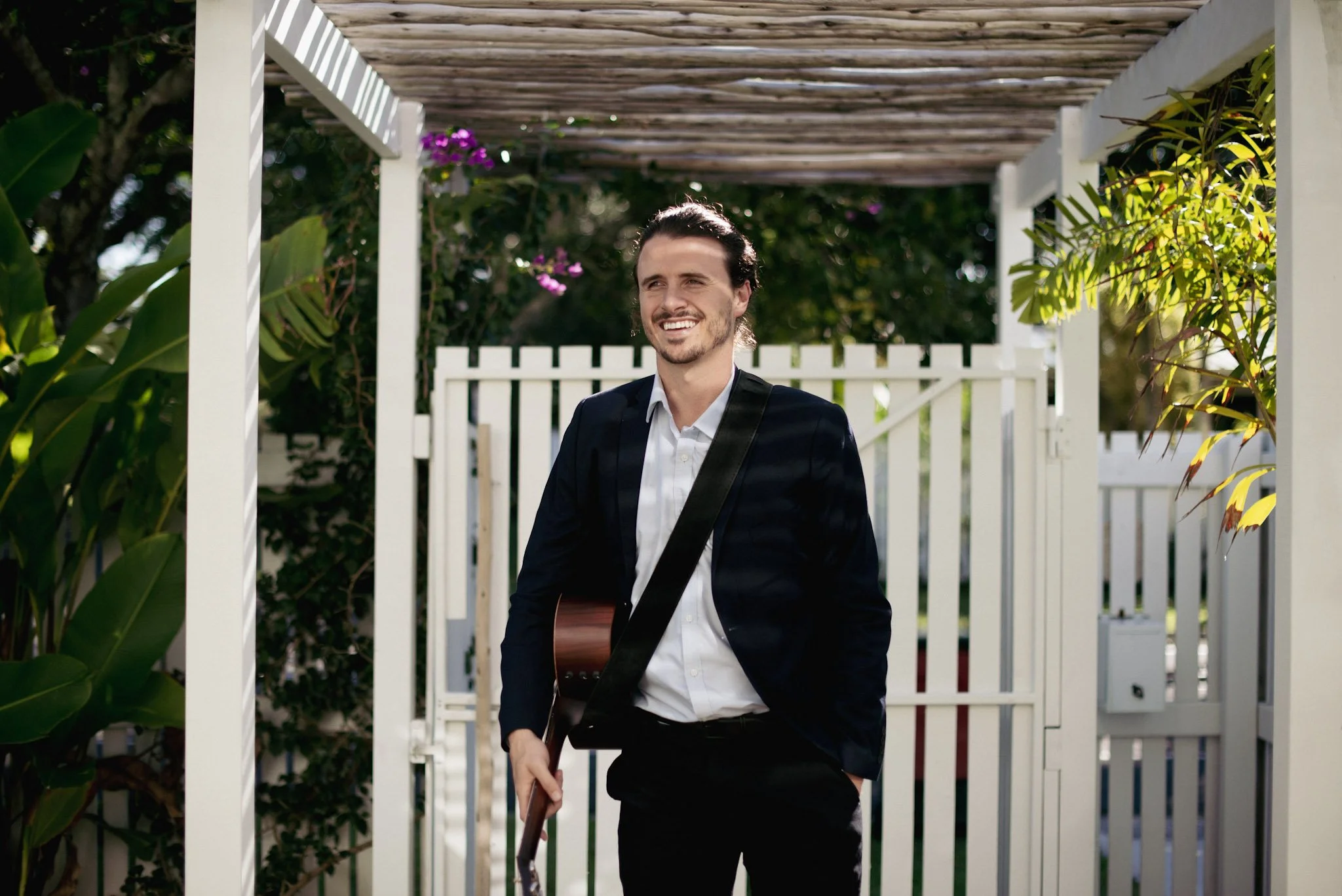 A man with dark hair and a beard, dressed in a white shirt and black blazer, stands outdoors holding a guitar. He is smiling and appears happy. He is framed by a white fence and greenery, with pink flowers and trees in the background.
